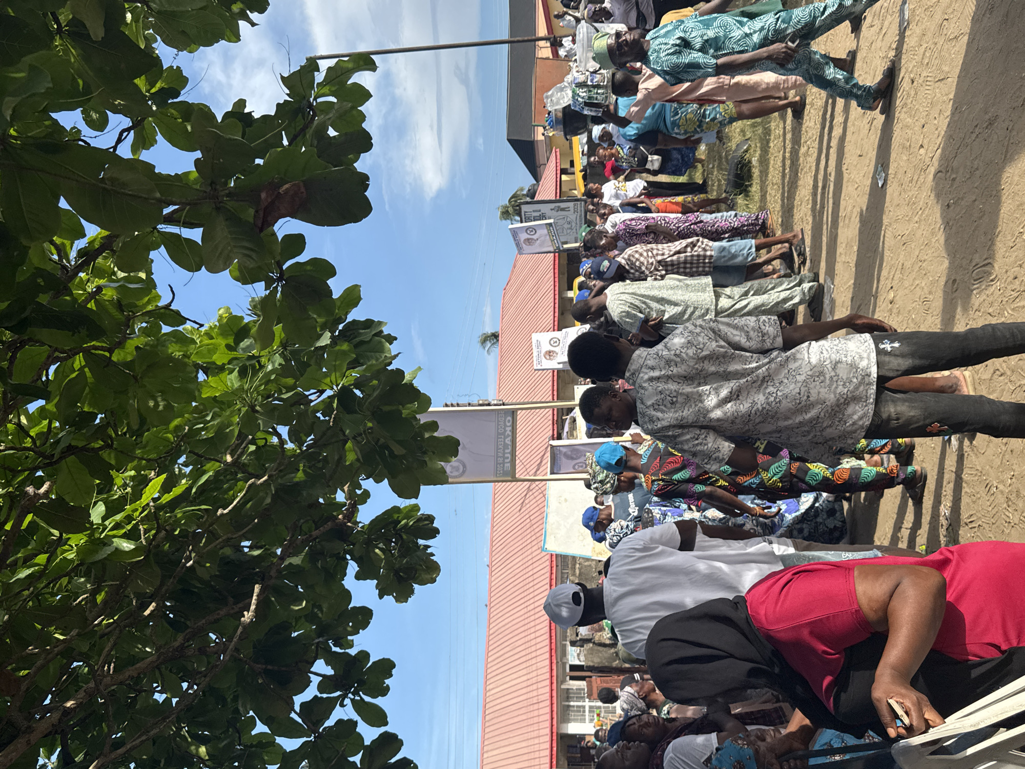 In Badagry, Nigeria, a large group of people are gathered outside a building with a red roof. They are standing in rows and facing towards the building. Some individuals are holding signs with what appears to be political images on them. The scene is outdoors, under a partly cloudy sky, and some trees are visible in the foreground. The environment suggests a community event or political gathering.