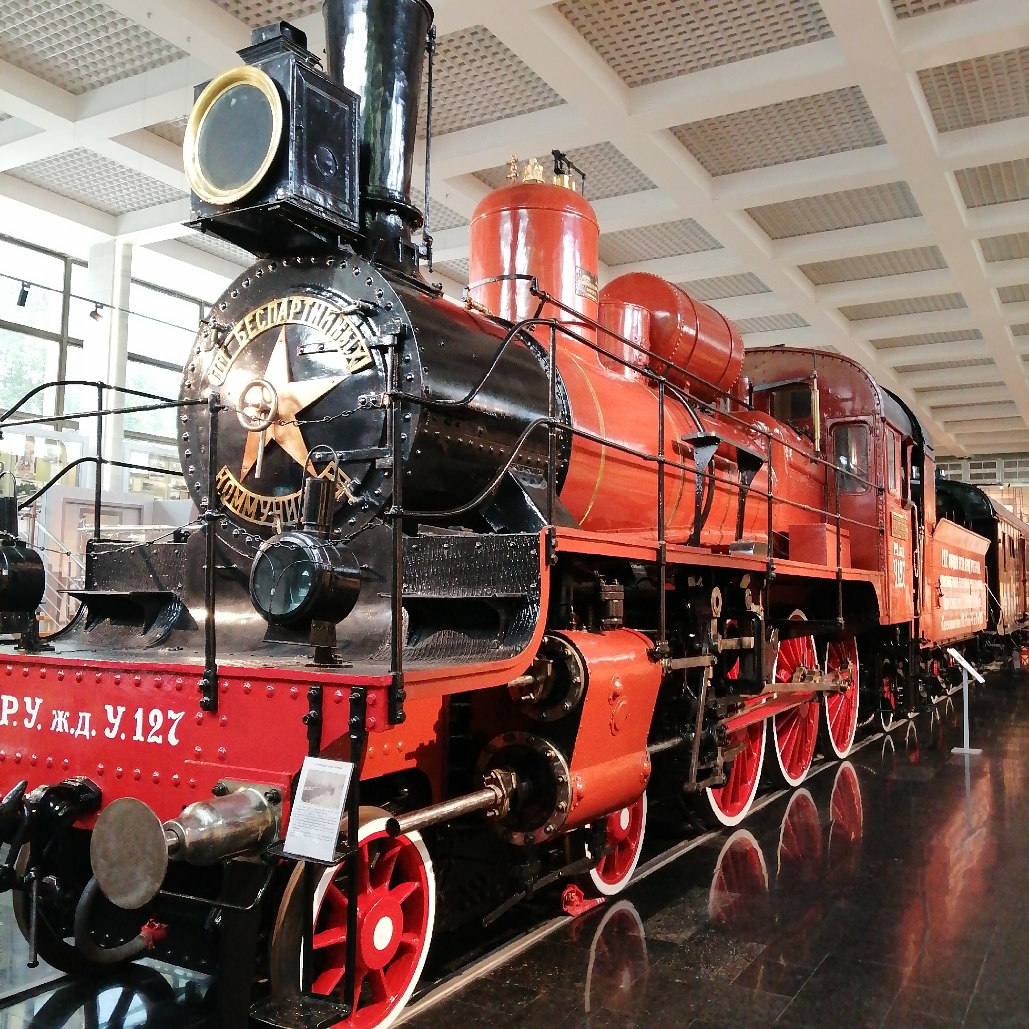 This image shows the Soviet locomotive U-127, displayed at the Moscow Railway Museum in Russia. The train is painted in red and black, with red wheels. White Russian lettering on the red portion of the train spells out “Р.У. Ж.Д. У.127.” The front of the train features a large gold star. The museum has a modern, grid-like ceiling. The floor is a highly polished, reflective dark marble. The locomotive is positioned on tracks within the museum setting.