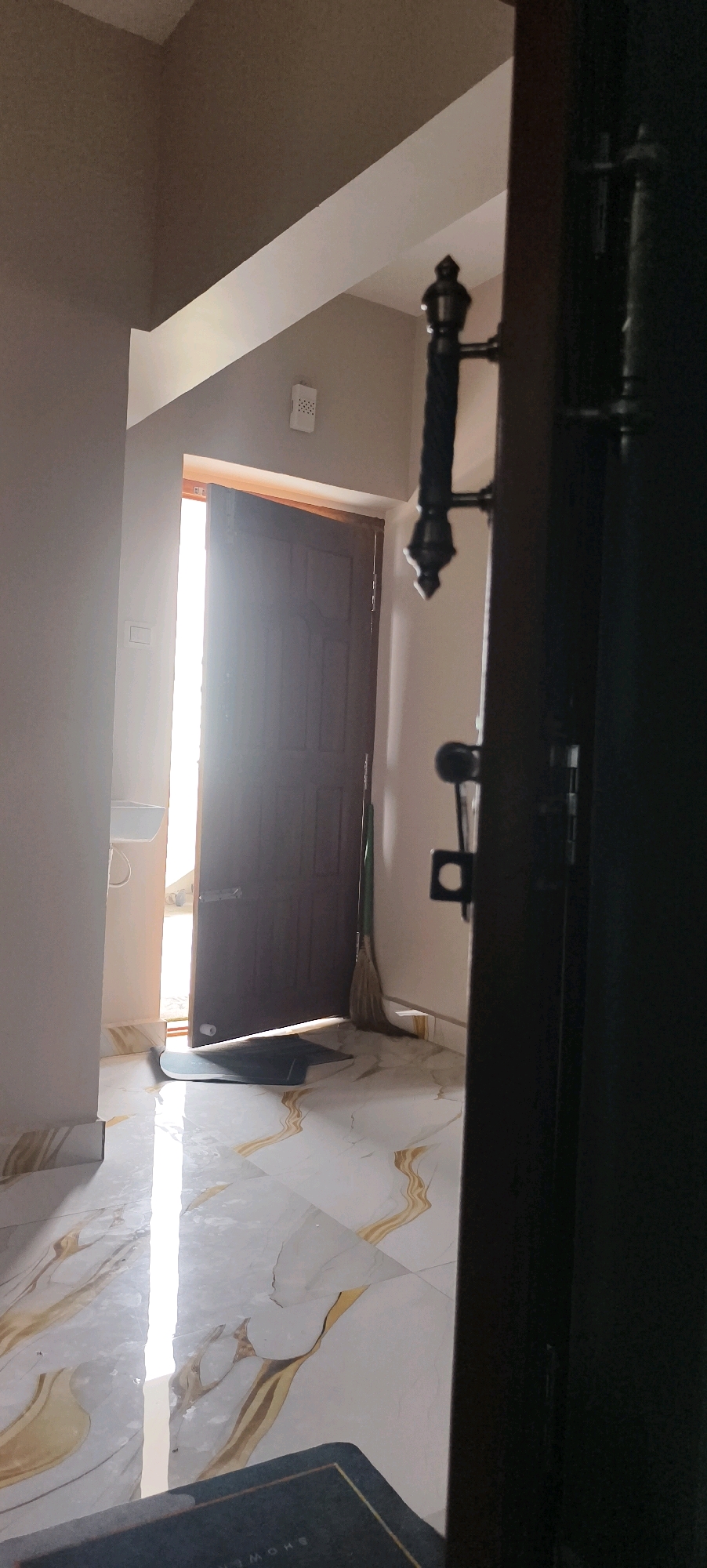 A dimly lit interior hallway is visible, with a dark-colored door partially open to a brightly lit exterior. The floor is tiled with a marble pattern featuring gold veining. To the left, a white sink is mounted on the wall. A broom leans against the open door. In the foreground, a dark doormat is partially visible on the floor. The scene appears to be located in Kundrathur, India.