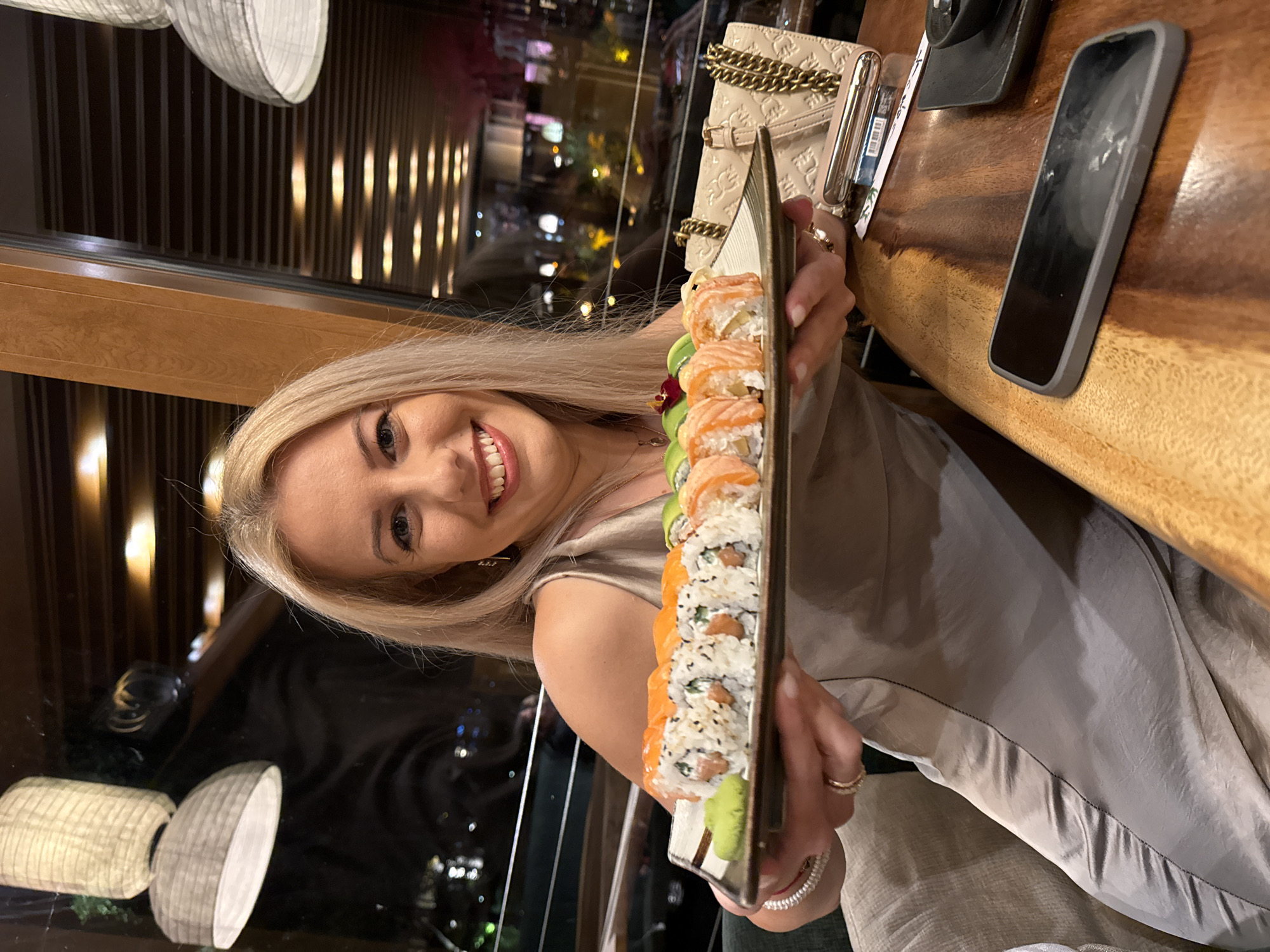 A woman with blonde hair is holding a platter of sushi in a restaurant setting, likely in Sofia, Bulgaria, given the context of the request. She is smiling and facing the camera. The platter contains various types of sushi rolls. A purse with a gold chain strap and a smartphone are visible on a wooden table next to her. The background shows an interior with wooden beams and large windows, implying a city view with lights at night.