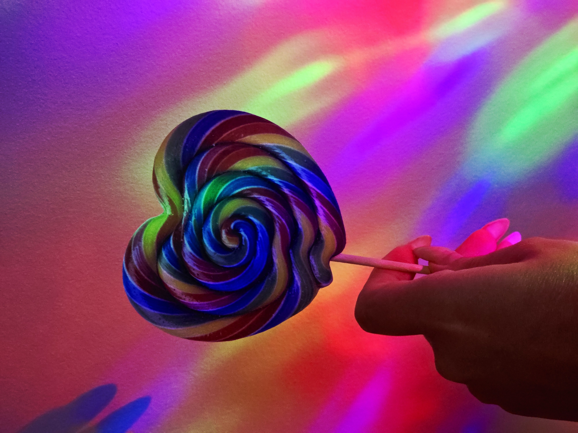 A hand holds a heart-shaped, multi-colored lollipop in front of a background illuminated by a spectrum of vibrant colors. The lollipop's swirls include shades of blue, yellow, red, and green. The hand is partially visible and seems to be holding the lollipop stick between fingers. The background transitions from red to pink, purple, and green, with streaks of light adding depth. The scene is brightly lit with neon hues.