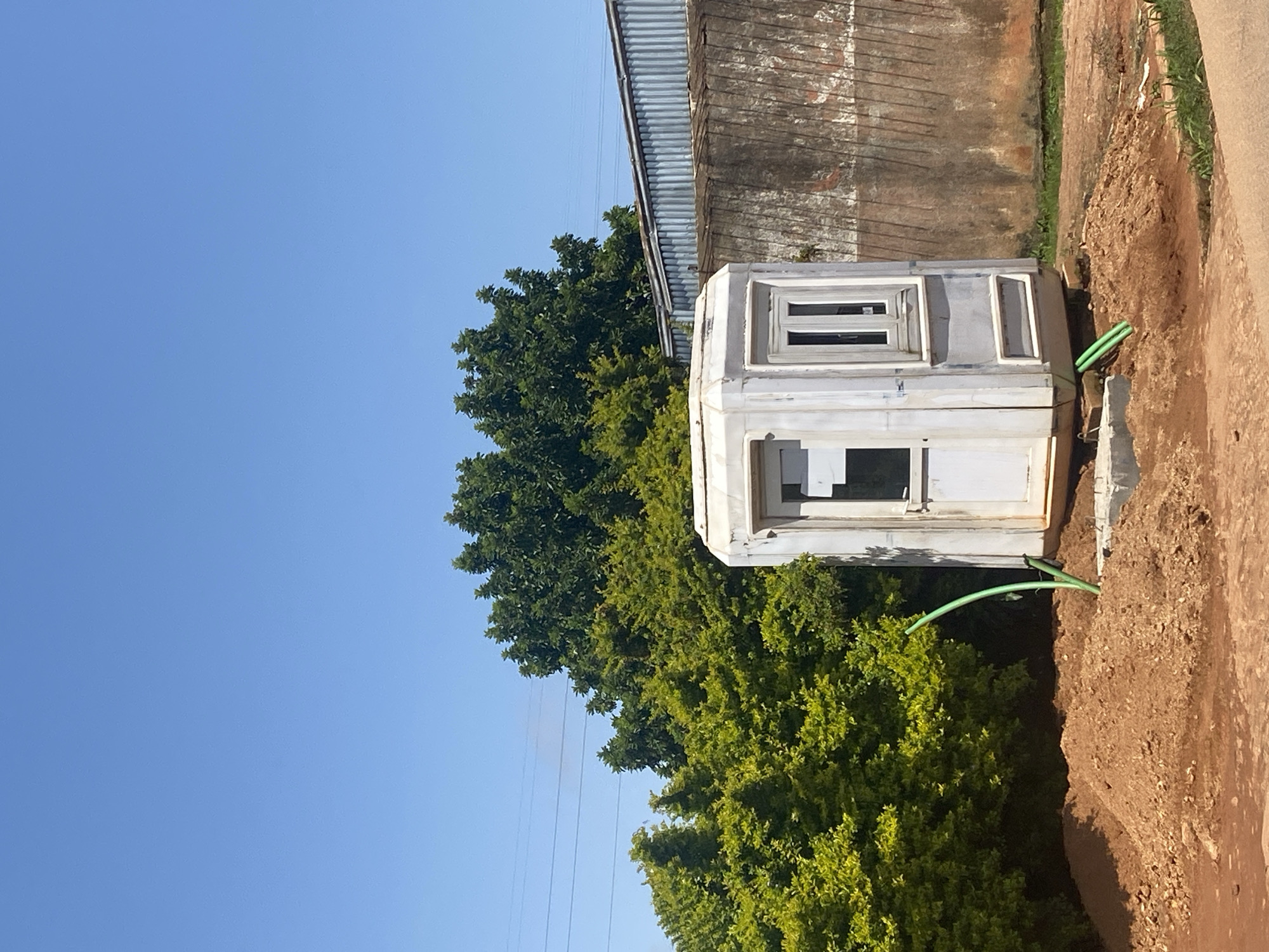 An outdoor shot shows a small, white, octagonal guardhouse with a single door and a two-pane window. The guardhouse is situated on a mound of reddish-brown dirt, with a green hose extending from it and bending downwards into the dirt. Behind the guardhouse, lush green trees create a dense backdrop, with some visible power lines overhead. To the right of the guardhouse, a corrugated metal fence runs horizontally, adjacent to a weathered, textured wall. The scene is set against a clear blue sky.