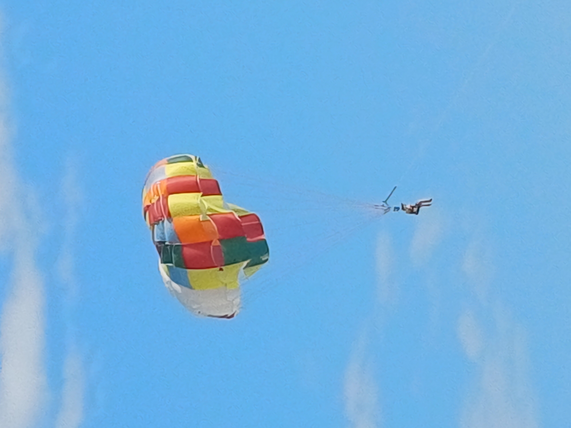 In a clear blue sky, a person is parasailing, suspended beneath a colorful parachute. The parachute is a vibrant patchwork of red, yellow, orange, green, and white squares, with the white section appearing to be the underside. The person, wearing what looks like a dark outfit, is positioned on a tow bar or harness, their legs extended as they ride the wind. The scene suggests an activity taking place on a sunny day, likely over water given the nature of parasailing, though no water is visible in this frame. The location is specified as Sochi, Russia. There is no visible text in the image. The overall impression is one of outdoor recreation and adventure.