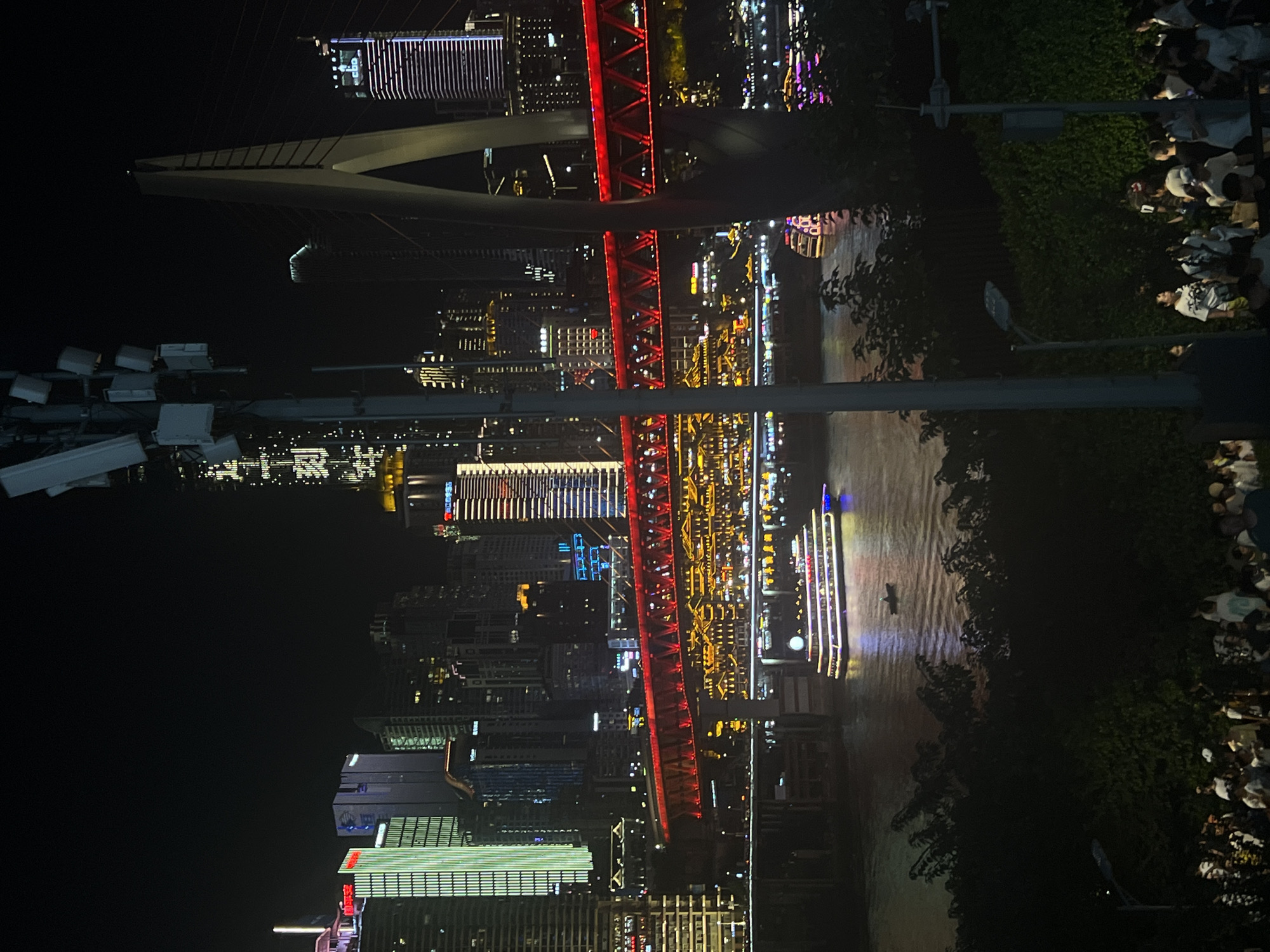 This image captures a nighttime cityscape along the Yangtze River in Jiangbeicheng Subdistrict, Chongqing, China. A brightly lit, multi-tiered bridge, illuminated with red and yellow lights, spans across the water. Several illuminated buildings, some with distinctive modern architecture and others with traditional Chinese-style roofs, form the urban skyline. A large passenger boat, adorned with colorful lights, is navigating the river. In the foreground, a crowd of people is gathered, appearing to be observing the illuminated scenery. The environment is dark, typical of nighttime, with artificial lights dominating the scene.