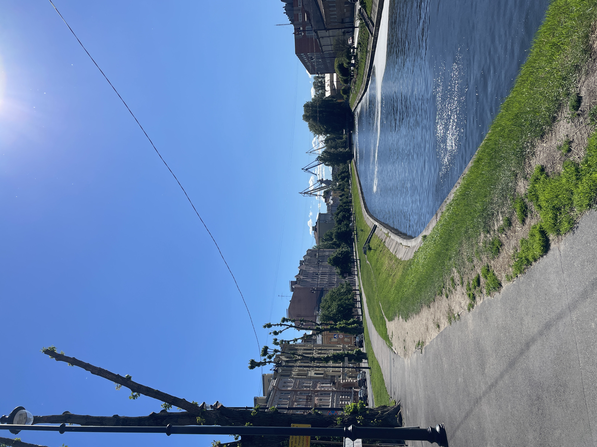 The image depicts a view of the Griboyedov Canal in Saint Petersburg, Russia, on a sunny day. A paved walkway lines the left bank of the canal, bordered by a grassy slope and trees. Across the canal, several multi-story buildings with distinct architectural styles are visible. In the distance, industrial cranes stand against the clear blue sky, suggesting the presence of a port or industrial area. The water of the canal reflects the sunlight, creating a shimmering effect. The scene is devoid of people, and the overall impression is one of urban tranquility.