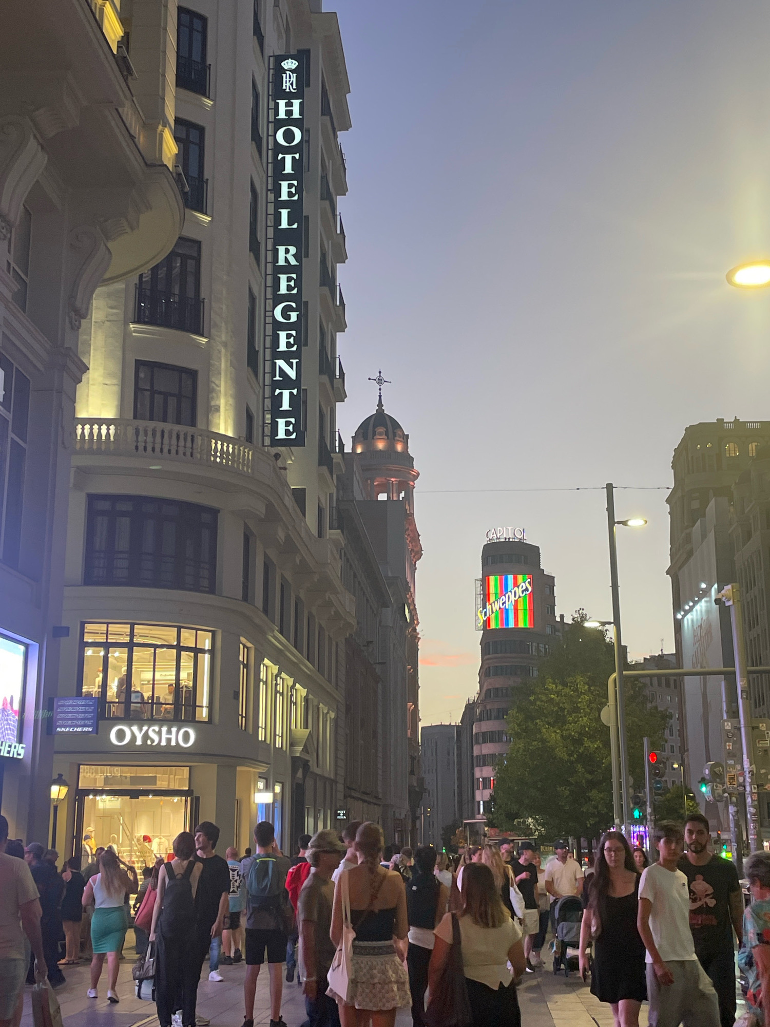 The image shows a street scene in Madrid, Spain, likely Gran Vía, in the evening. A prominent vertical sign on the left reads "HOTEL REGENTE" with a crown logo above it. Below the hotel sign, the OYSHO store is visible with its illuminated interior and storefront. A crowd of people is walking on the sidewalk. In the background, a distinctive building with a large "CAPITOL" sign and a "Schweppes" advertisement featuring a colorful screen is visible. The sky is a soft gradient of purple and orange, indicating twilight. Streetlights and building lights illuminate the scene.