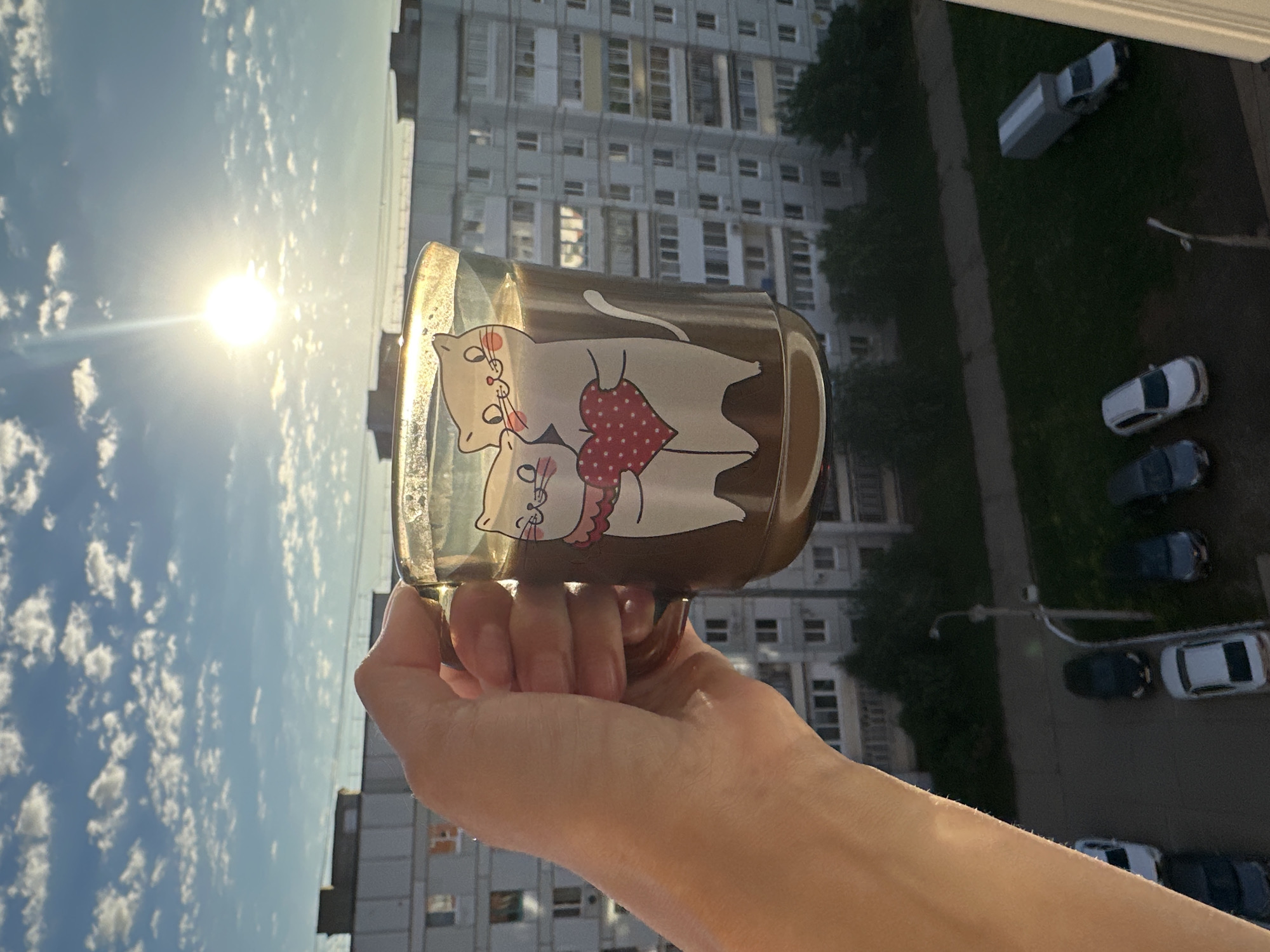 An individual's hand holds a clear glass mug filled with a brown liquid, possibly coffee or tea, adorned with a decal of two white cats embracing a red heart. The mug is positioned in front of a large, multi-story apartment building under a bright, sunny sky with scattered clouds. The scene appears to be from a balcony or window overlooking an urban environment, with cars parked below on a street and a patch of green grass. The sun is high in the sky, casting a strong glare. The architectural style of the apartment building suggests it is located in Chaikovsky, Russia, a city known for its residential high-rises. The overall impression is a casual, domestic moment captured outdoors, likely in the late afternoon or early morning given the angle of the sun.