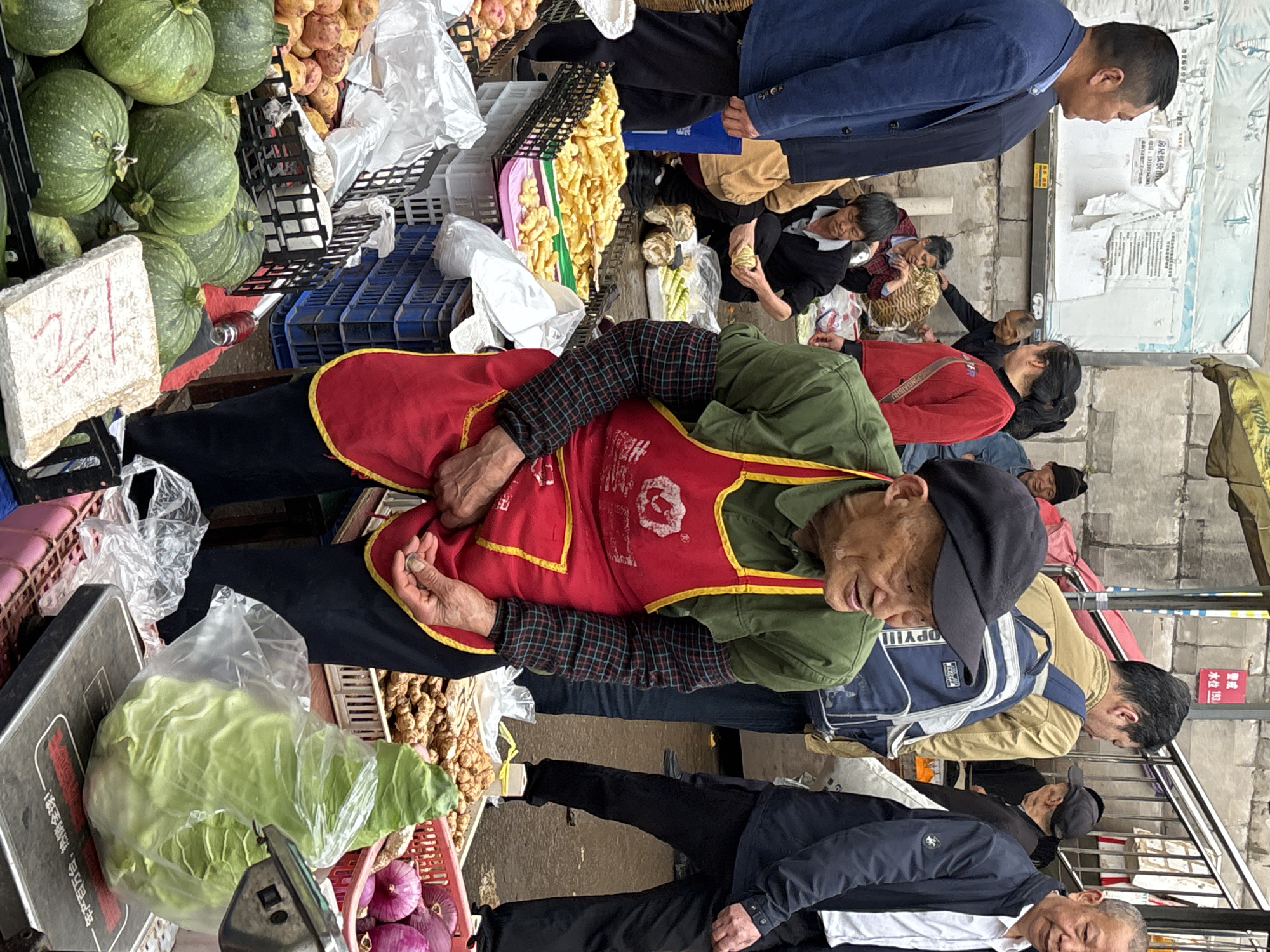 The image shows a bustling street market scene in Jiangjin, China. Several people are present, including vendors and shoppers. One vendor, wearing a red apron over an olive green shirt and a black flat cap, is counting money while sitting behind a display of various produce. The produce includes green pumpkins, potatoes, ginger, cabbage in a plastic bag, and red onions in a pink plastic basket. There's a sign indicating the price of an item, with the number "21" prominently displayed. Shoppers are perusing the stalls, and some appear to be engaged in transactions. The background includes a concrete wall with posters and signs in Chinese. The setting is urban, with an atmosphere of activity and commerce.
