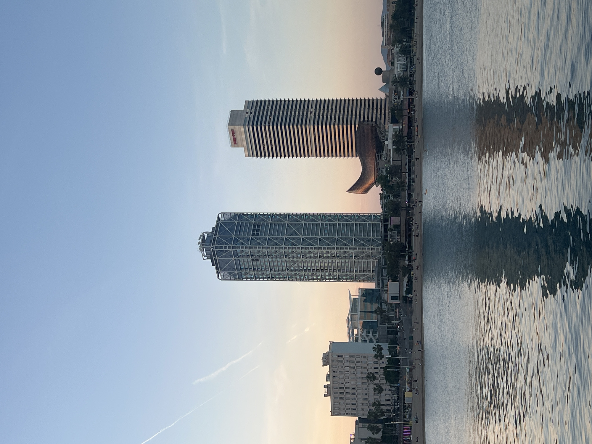 The image shows the Barcelona coastline featuring two iconic skyscrapers, Hotel Arts and Torre Mapfre. The buildings stand side-by-side against a light blue and orange-tinted sky. To the right of the buildings is a coastline. The water creates distorted reflections of the beachfront.