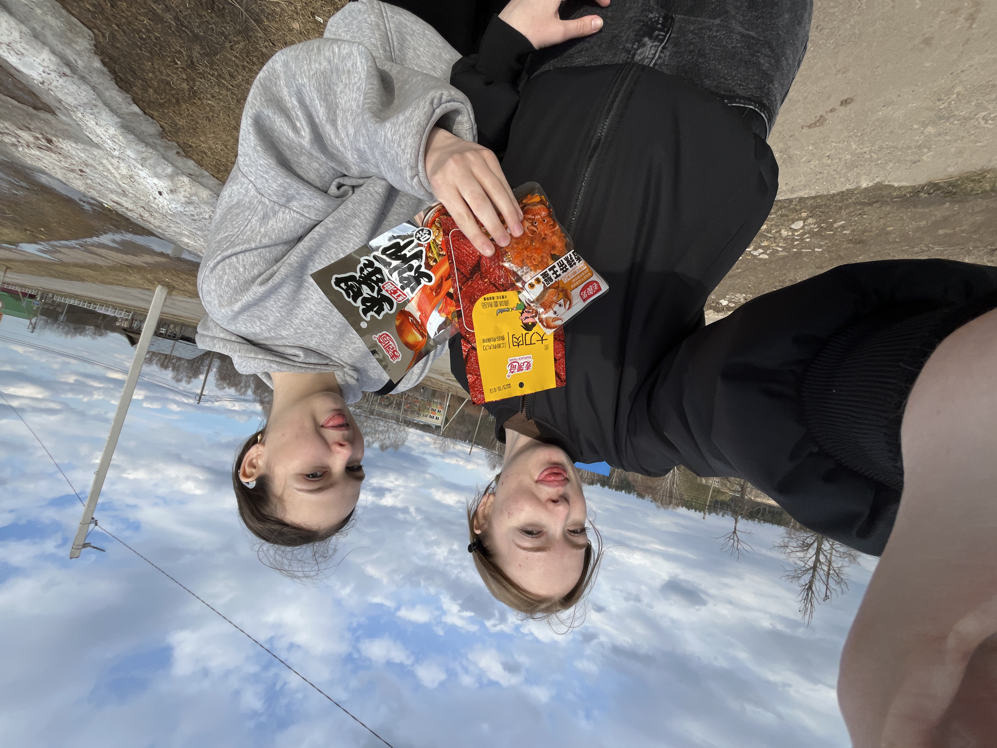 The image, taken in Сосново, Russia, captures two young people posing for a casual outdoor selfie. The original orientation of the image is vertical, rotated 90 degrees counter-clockwise, making the subjects appear upside down relative to the ground. When viewed upright, two individuals, likely young women, are seen from the chest up. The person on the left wears a grey hoodie, while the person on the right is in a black jacket and dark clothing. Both are playfully sticking out their tongues.
They are holding two distinct snack packages. The package on the left is black and silver, featuring Chinese characters reading "毛俊男" (Mao Jun Nan), "香辣帝王蟹" (Spicy King Crab), and the brand "麦凯奇" (Makala Foods). The second package, held by the person on the right, is primarily yellow and red, displaying "大刀肉" (Big Knife Meat), along with the phrase "江湖有大刀 食品有麻辣" (There's a big knife in the Jianghu, and spicy food). This package also shows the brand "麦凯奇" and a date "2005/10/013".
The setting appears to be an open, semi-rural area, possibly a roadside or path. The ground shows patches of melting snow and ice alongside dirt or gravel, indicative of early spring weather. Bare trees are visible in the background, along with distant buildings and a utility pole with wires stretching across a cloudy but bright daytime sky. The overall mood conveyed is lighthearted, candid, and friendly, capturing a moment of shared enjoyment of snacks outdoors.