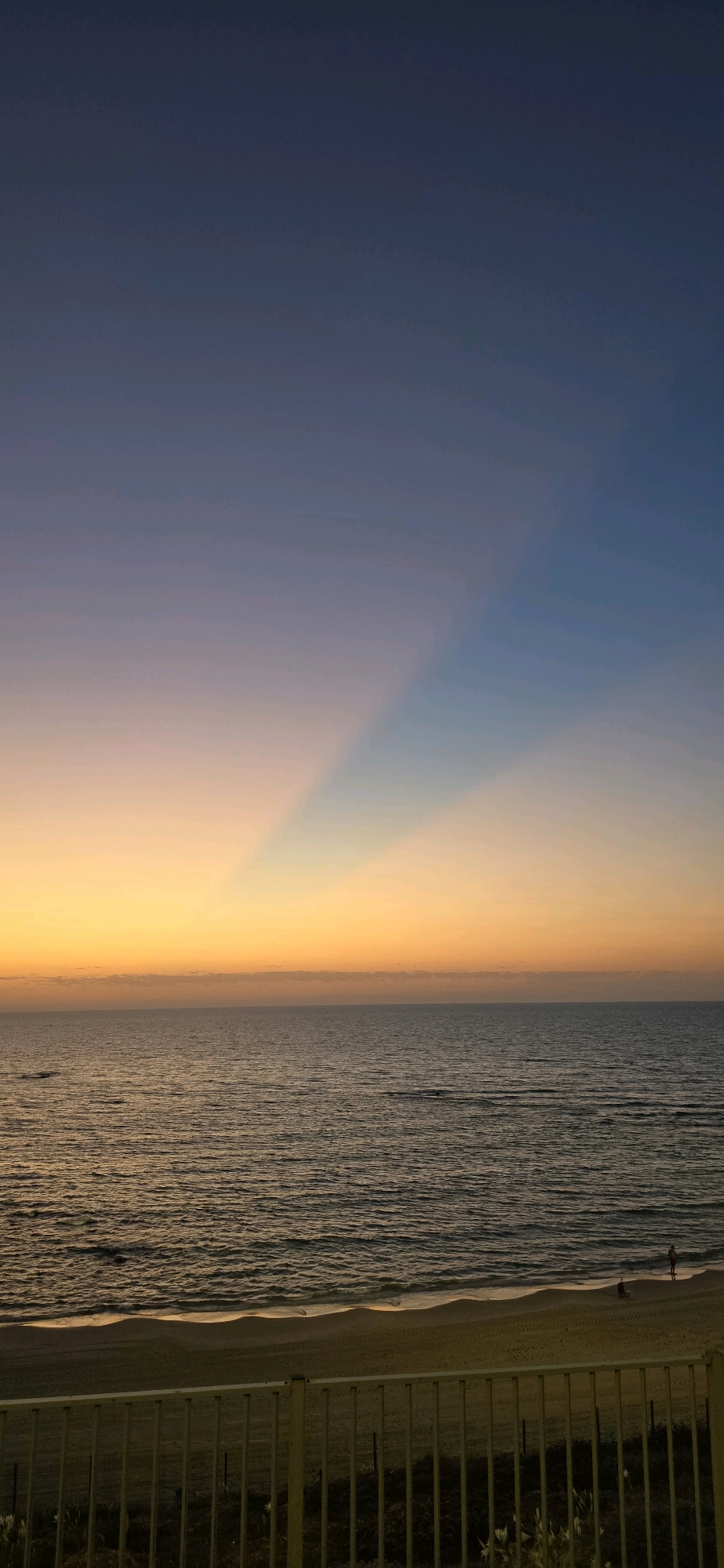 The image shows a serene sunset over the Mediterranean Sea at Bat Yam, Israel. The sky transitions from a deep blue at the top to a soft orange and yellow near the horizon, with distinct sunbeams fanning out. The calm sea reflects the warm hues of the sky. A sandy beach is visible in the foreground, with a few people walking along the shoreline. A yellow railing runs along the bottom of the image, separating the viewer from the beach. The overall atmosphere is peaceful and tranquil.
