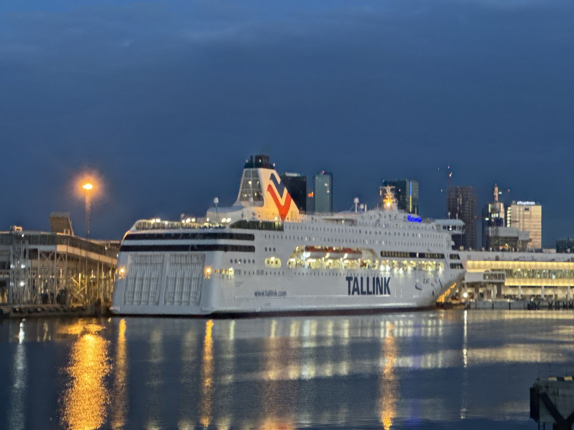 The image depicts a large passenger ferry docked at a harbor in Tallinn, Estonia, during the twilight hours. The ferry, identified by the "TALLINK" logo on its side, is illuminated, with lights visible through its windows and on the deck. The ferry's hull is white with a red waterline. The ferry's stern is facing the viewer, and its broad side stretches across the frame.
Behind the ferry, the city skyline of Tallinn is visible, featuring several modern buildings with illuminated windows and some construction cranes. A prominent streetlight with a bright orange glow is positioned to the left of the ferry, casting reflections on the water. The water itself is dark, reflecting the lights from the ferry and the city. The sky is a deep blue, characteristic of dusk or dawn. The scene appears to be at a port terminal, with structures and walkways associated with ferry operations visible along the dock. There are no people clearly visible in the image. The overall impression is of a quiet, active port at the edge of a city.