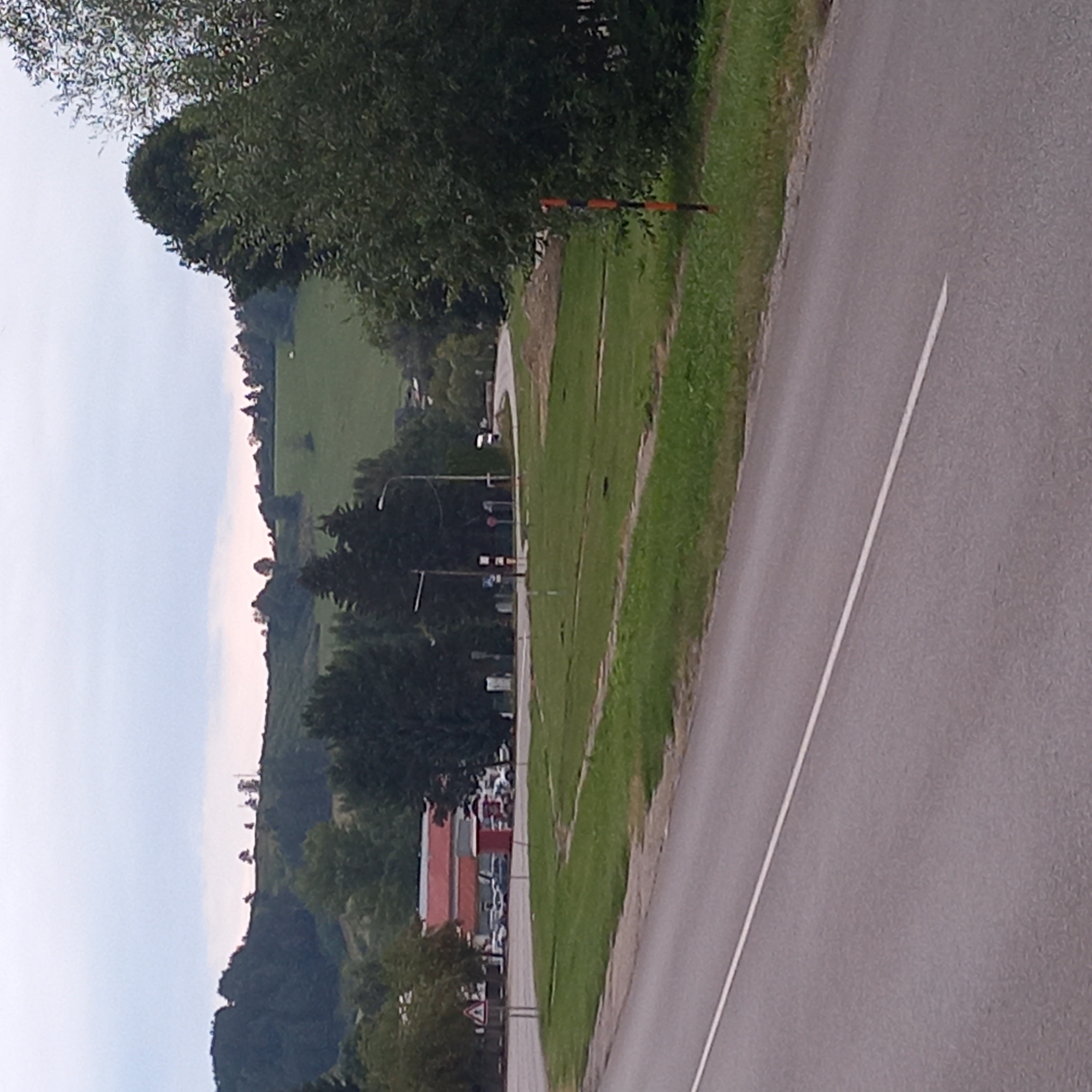 The image shows a roadside scene in Dolný Kubín, Slovakia. A paved road with a white line marking runs diagonally from the bottom left to the top right of the frame. To the right of the road is a grassy area with some trees and foliage. In the mid-ground, there are several buildings with red roofs, appearing to be commercial structures, and a number of parked cars. Further in the background, a winding road leads into a landscape of rolling green hills covered with trees, with a cloudy sky overhead. A striped orange and black marker post is visible in the grass on the right side of the image.