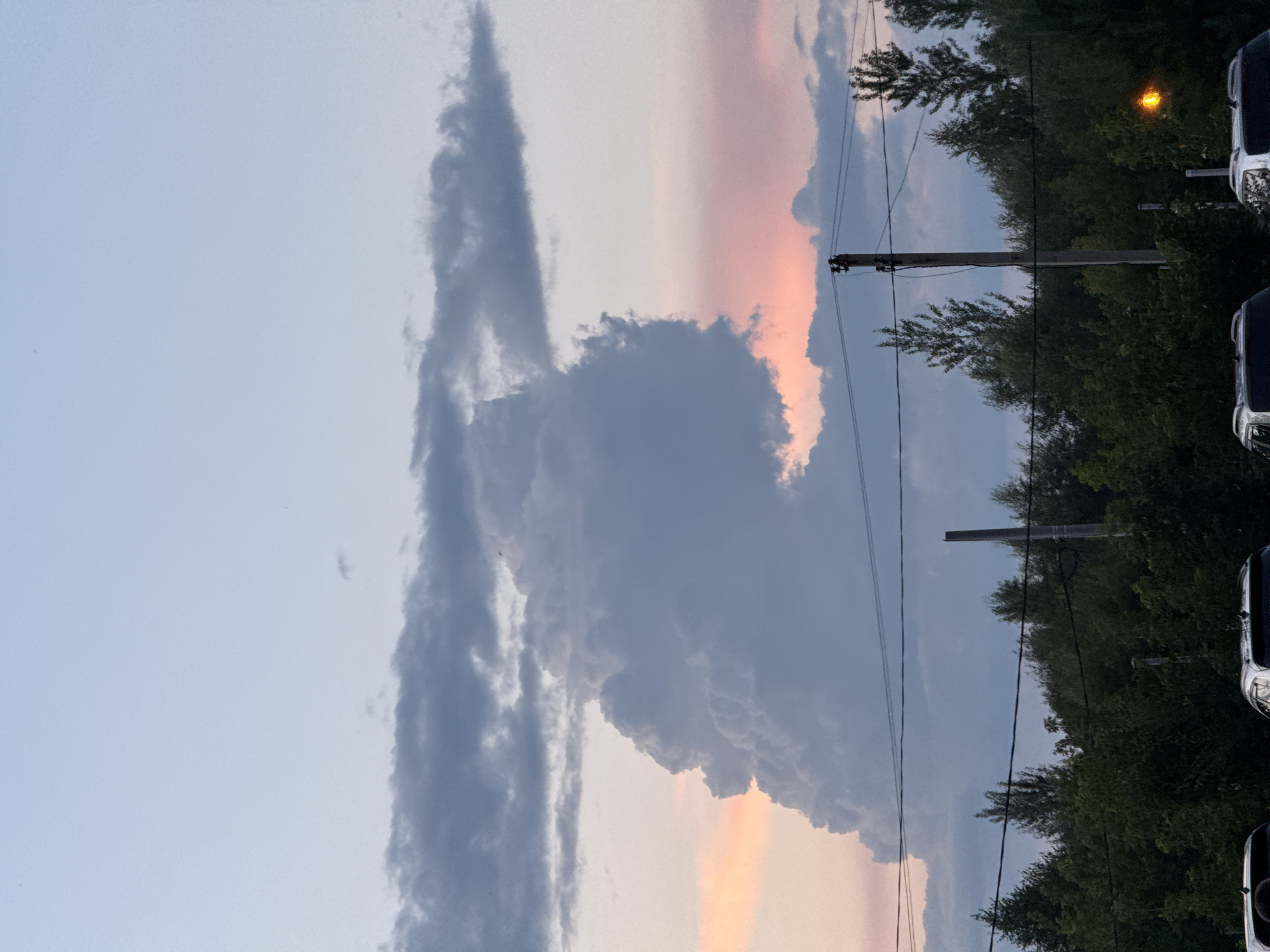 The image shows a dramatic sky with large, dark clouds dominating the center. To the left, there is a lighter pinkish hue, suggesting a sunset or sunrise. Below the sky, a line of dense green trees stretches across the frame, punctuated by utility poles and power lines. In the foreground, the tops of several cars are visible, parked side-by-side. The scene appears to be a street or parking area in the city of Otradny, Russia, at dusk.