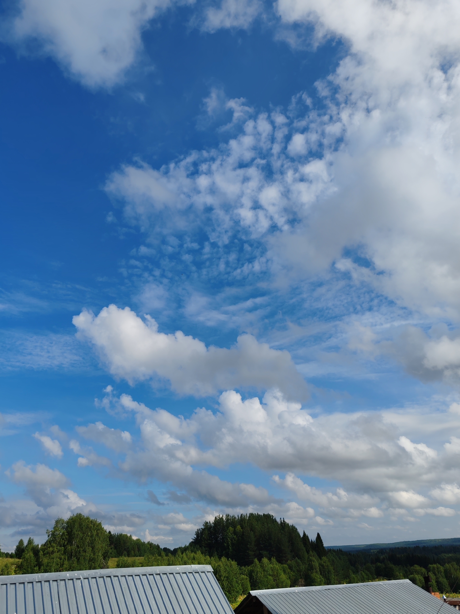 The image shows a wide expanse of a bright blue sky filled with various types of clouds, from wispy cirrus formations to larger, puffier cumulus clouds. The clouds are white and grey, with some areas of shadow indicating their depth. Below the sky, a green landscape unfolds, characterized by dense forests and rolling hills. In the foreground, the corrugated metal roof of a building is visible, extending across the bottom of the frame. The scene appears to be an outdoor view from a high vantage point, likely a rooftop or elevated area, within the city of Chukhlem, Russia. No people or specific actions are depicted.
