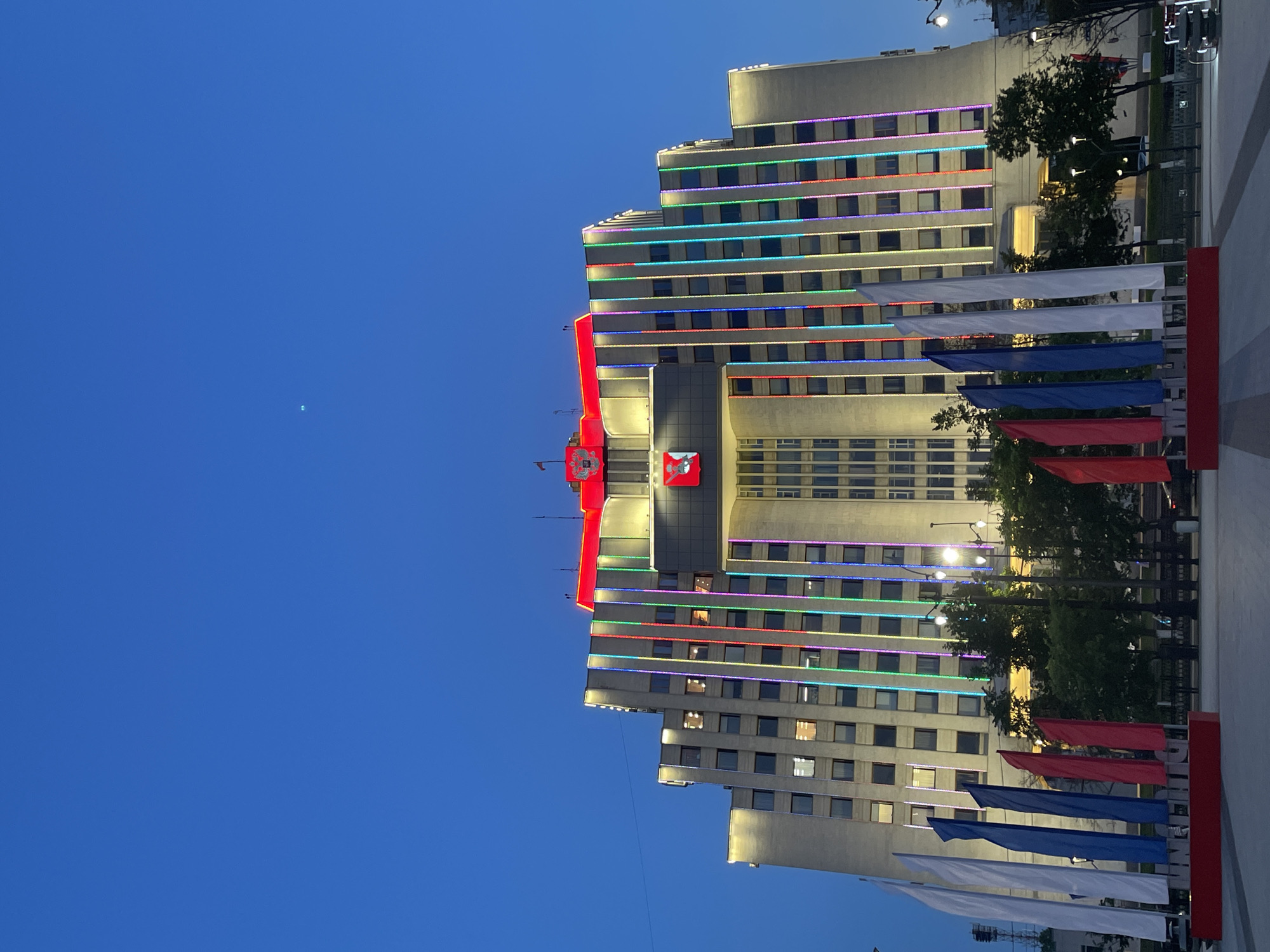 The image shows the Vologda City Duma building in Vologda, Russia, at dusk. The building is illuminated with colorful LED lights along its facade, with a red illuminated section at the top featuring the city's coat of arms. In the foreground, several flags are displayed, including white, blue, and red flags, arranged in front of a red base with white lettering. The sky is a deep blue, indicating the transition from day to night. Trees and streetlights are also visible in the background.