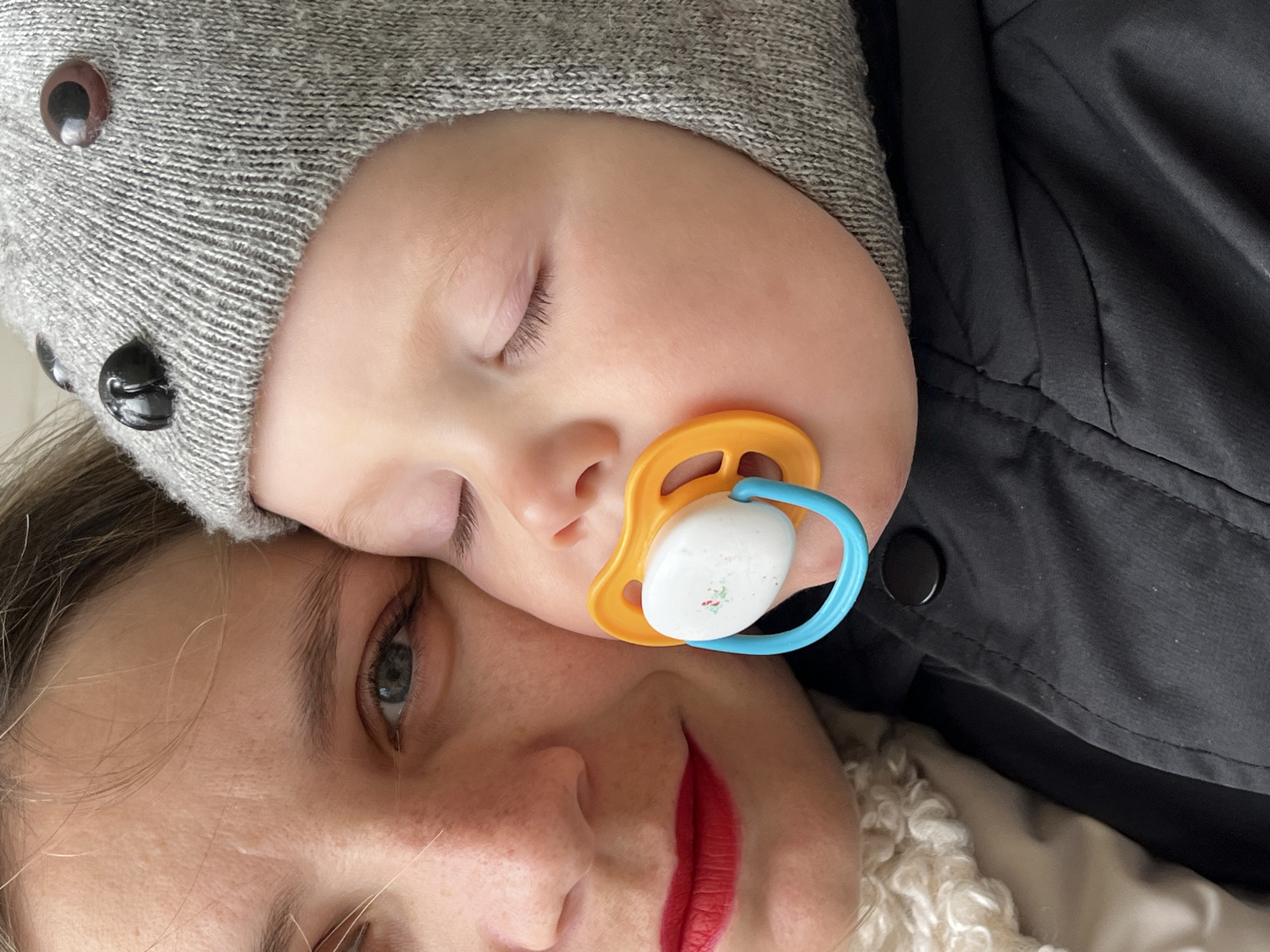 A close-up shot shows a baby with a gray knit hat with googly eyes on their head resting against a woman's face. The baby's eyes are closed and they have a pacifier in their mouth. The woman has fair skin and red lipstick. The background includes a black jacket and a white furry material. The image appears to be taken in an indoor setting. There is no contextual information in the image to connect it to Chaikovsky, Russia.