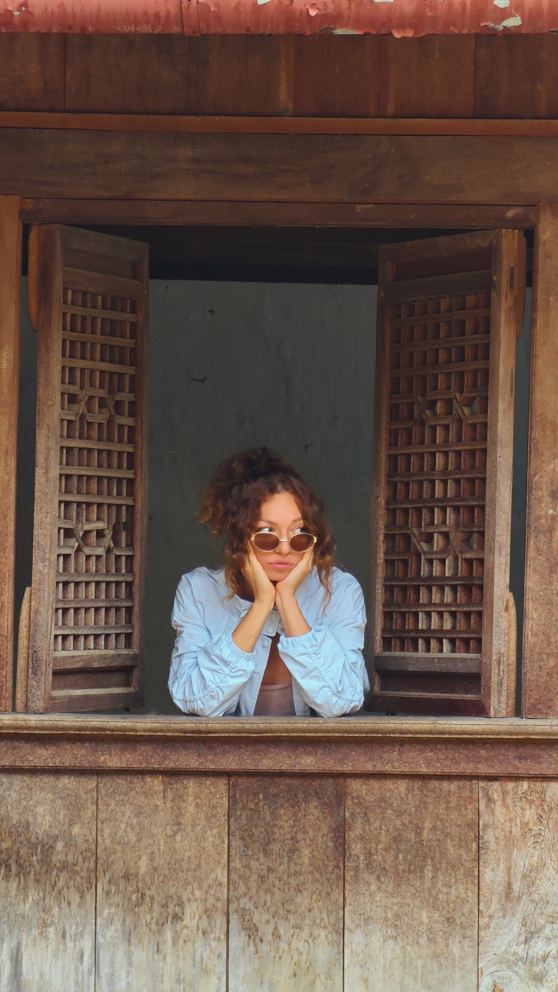 A medium shot depicts a woman seated in a traditional wooden window in Sà Phìn Commune, Vietnam. The woman has curly red hair, wears round sunglasses, and a light blue shirt. She rests her chin in her hands, gazing out the window. The window frame is dark wood, and the shutters are partly open, revealing a gray wall behind the woman. The window frame is textured, and the shutters have a square lattice pattern. The bottom part of the window has a panelled wood surface.