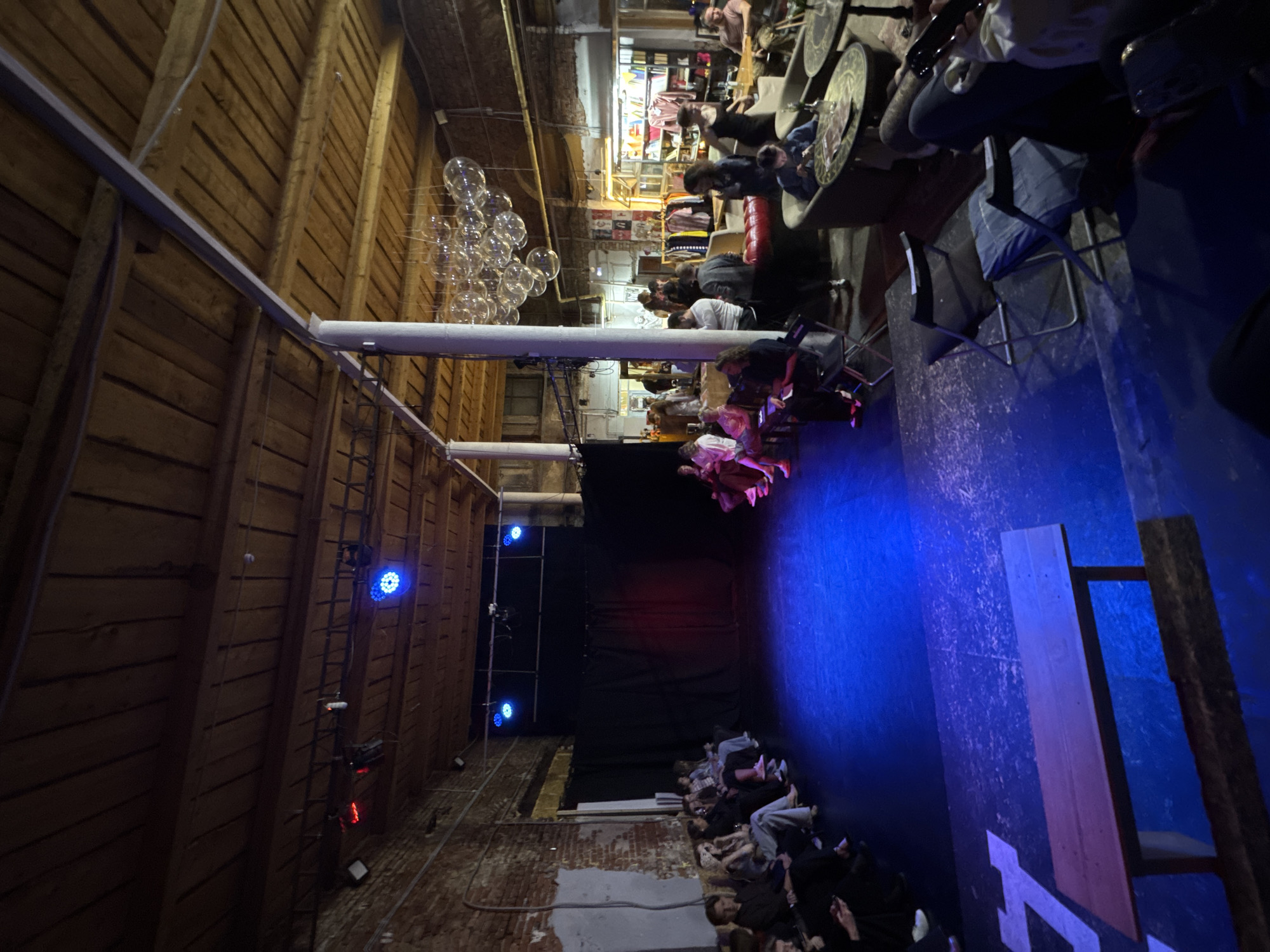 The image shows an indoor performance venue in Izhevsk, Russia. The space has a stage illuminated with blue lighting, and a seated audience is visible on both sides of the stage. The venue appears to be in a refurbished industrial building, with exposed brick walls and wooden ceilings. Decorations include spherical hanging lights and mounted lighting fixtures. Some audience members are seated at tables, while others are in rows of chairs. The audience seems to be watching a performance on the stage.