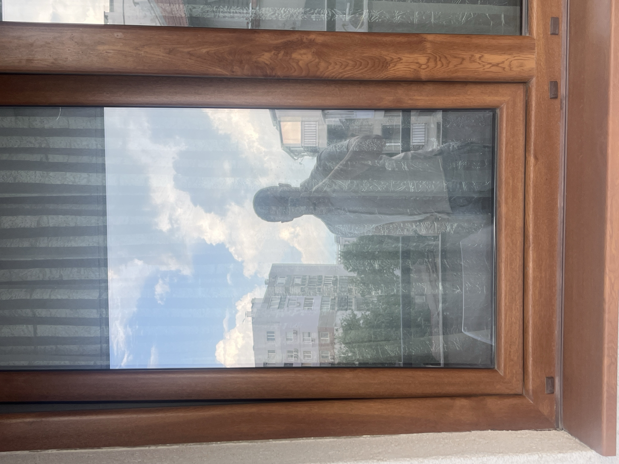 The image shows a reflection in a window, framed by a brown wooden window frame. The reflection contains a view of a city landscape with a statue visible on the right. A building with multiple floors and windows is also visible. The presence of a monument to the late Bulgarian communist and anti-fascist politician Georgi Dimitrov suggests that this scene is located in Varna, Bulgaria.