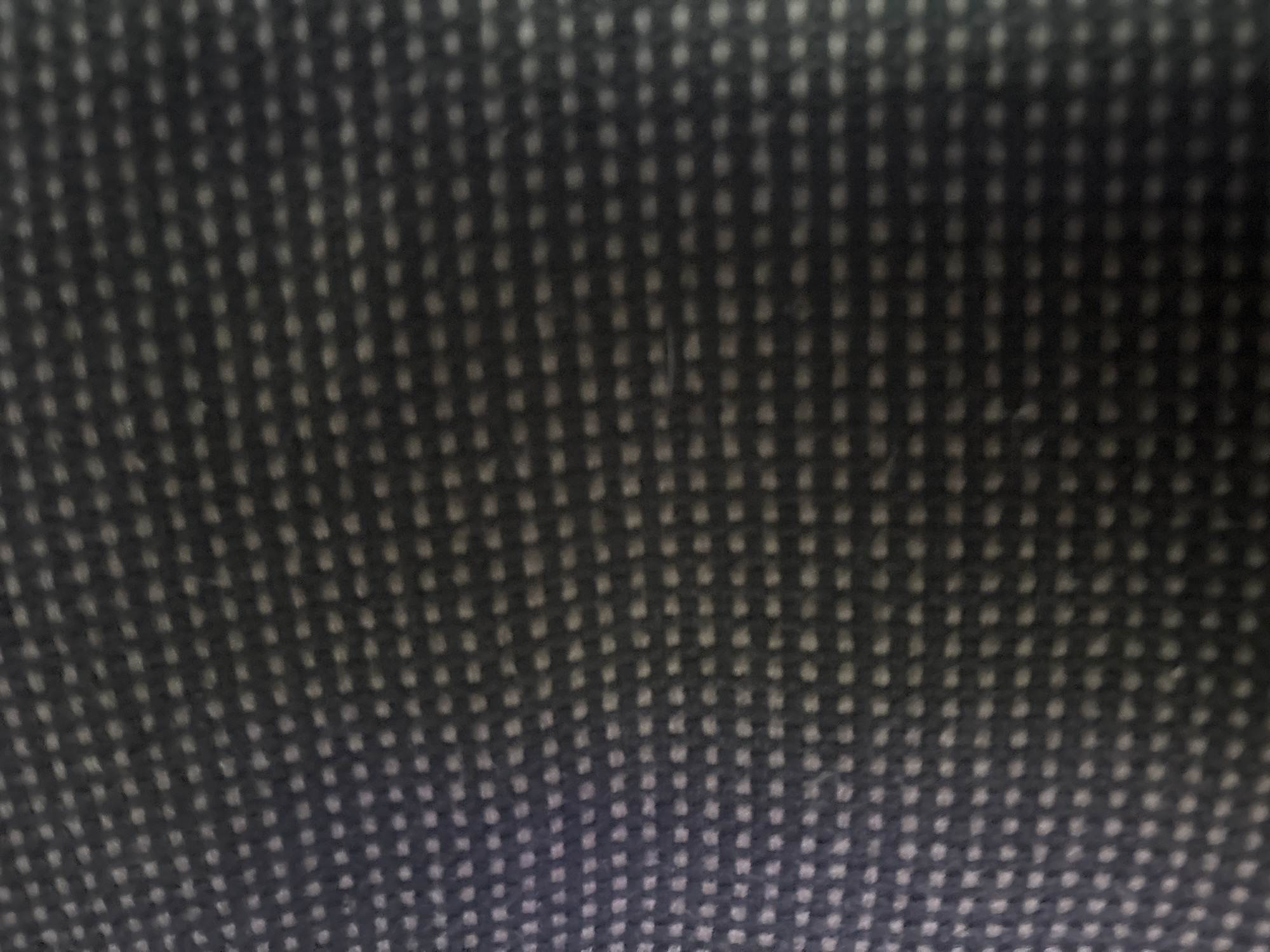 The image is a close-up, overhead shot of a dark blue, woven fabric with a dotted pattern of lighter blue or gray threads. The fabric appears to be textured and possibly a carpet or upholstery. The lighting is dim, creating shadows and highlighting the texture of the material. There are no discernible objects, people, or specific environmental features.