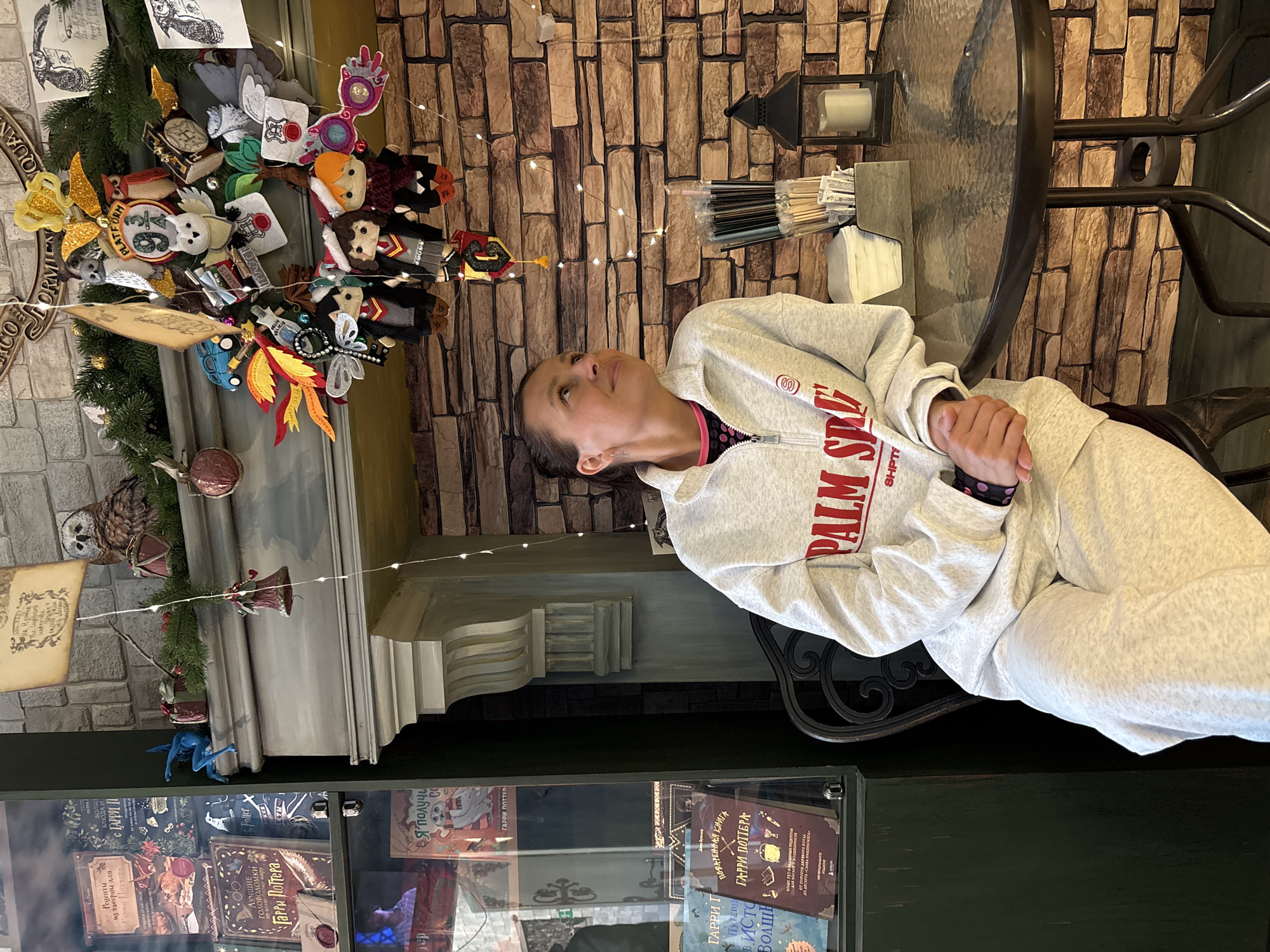 The image depicts a woman sitting at a round, glass-topped table in what appears to be a Harry Potter-themed establishment in Kazan, Russia. The woman, wearing a light grey tracksuit with the word "PALM SPR" written on it in red, is looking upwards and to the left.
Behind her is a wall with a brick-like pattern adorned with fairy lights. A lantern sits on the table beside her. To the left is a display of Harry Potter-themed ornaments, including stuffed figures and other decorative items attached to a faux evergreen branch. Below the ornaments is a glass display case containing various editions of Harry Potter books in Russian. The OCR provided with the image lists titles such as "Рецепты из таверны для Гарри Поттера" and "ГАРРИ ПОТТЕРА."
Overall, the scene suggests a casual, themed environment.