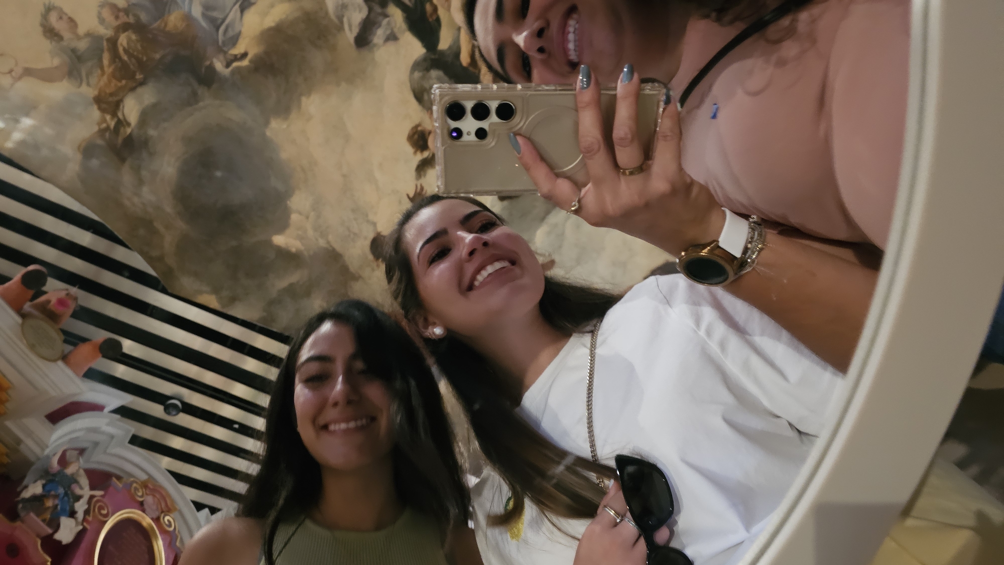 Three smiling women are reflected in a mirror. The woman closest to the camera holds a cell phone, presumably to take a photo. She wears a watch on her left wrist and has a ring on her left hand. The other two women are positioned to her left. The woman in the mirror wears a pink shirt, while the other two women wear a white t-shirt and a green tank top. Behind the women are black and white stripes, an ornate carousel feature, and a painted ceiling. The background details indicate that they are at the Grand Canal Shoppes in the Venetian Resort Hotel Casino, located in San Bernardino Tlaxcalancingo, Mexico.