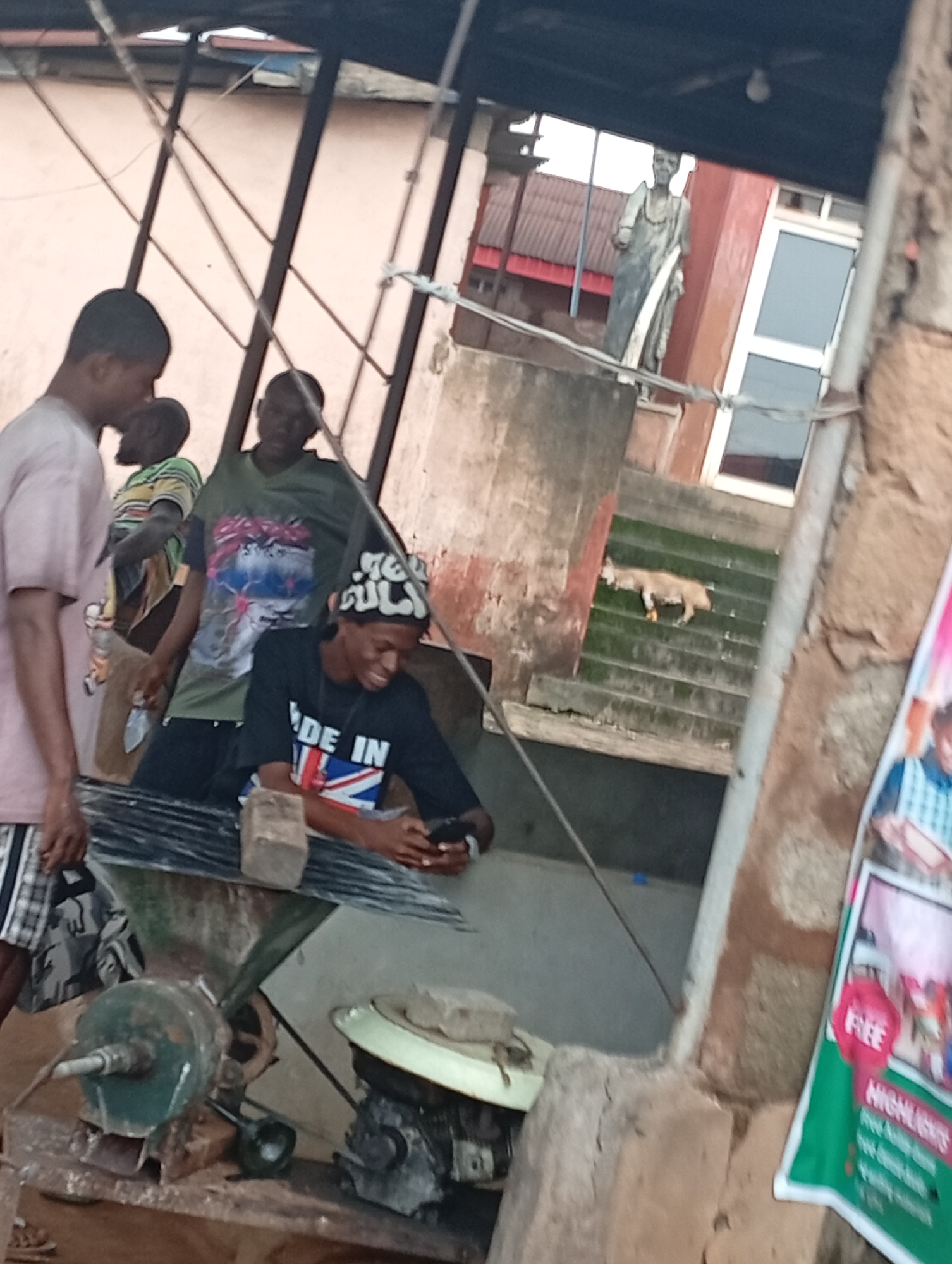 The image depicts an outdoor scene in Nsugbe, Nigeria. In the foreground, a young man wearing a black t-shirt with a Union Jack design and a patterned cap is looking at his phone and smiling. He is seated next to a metal contraption, possibly a grain mill, made of rusted metal parts and a green hopper. Beside him, another young man in a green t-shirt with a graphic print is standing. To his left, a third young man, partially visible, is wearing a light-colored t-shirt and plaid shorts. In the background, a dog is seen on a set of green outdoor stairs. A statue of a robed figure stands on a higher level, and a red-roofed building is visible further back. A poster with the word "FREE" and "HIGHLIGHTS" is attached to a rough-textured wall on the right side of the frame. The overall environment appears to be a communal or work area, possibly on a street or in a yard, with a mix of utilitarian objects and architectural elements.