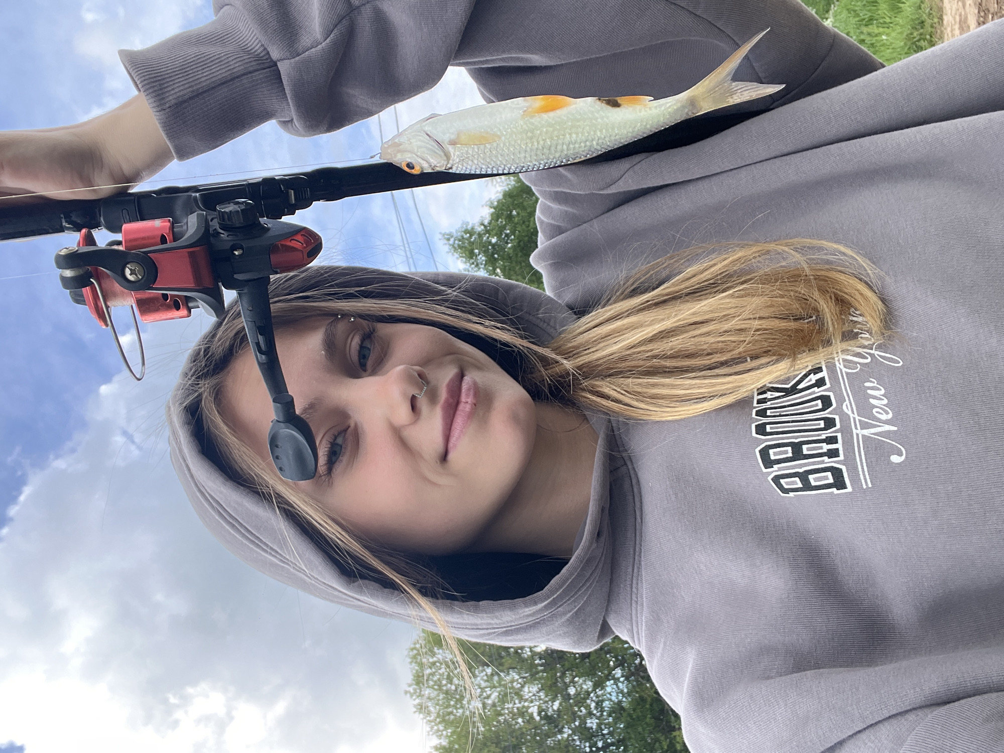 A person with light blonde hair is holding a fishing rod and a small fish. The person is wearing a gray hooded sweatshirt with "BROOKLYN New York" printed on it. The fishing rod has a red and black reel. The fish is silver with yellow markings. The background includes a cloudy sky, trees, and power lines. The location in the city of Фоки, Russia is not discernible from the given image.