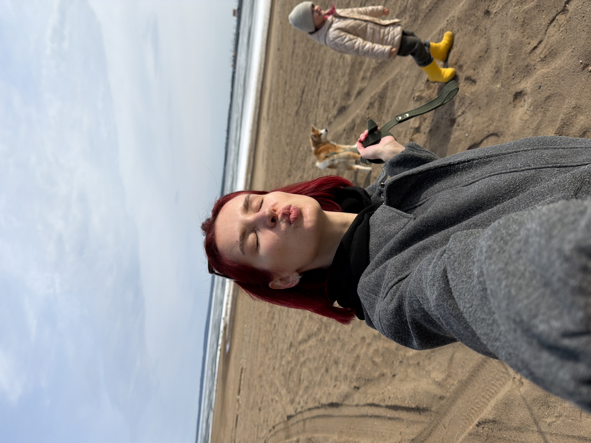 A woman with red hair is taking a selfie on a sandy beach. She is wearing a gray coat over a black hoodie and is holding a green dog leash. Her lips are pursed. In the background, a child in a beige jacket and yellow boots is walking away, and a dog is standing near the woman. The background also shows a body of water and a partly cloudy sky. The location is Noviy, Russia. The environment appears to be a beach, likely next to a large body of water, perhaps a lake or sea. There are tire tracks in the sand.