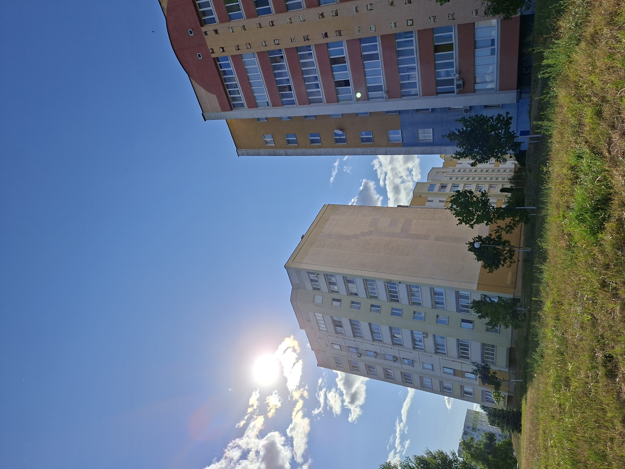 The image depicts a bright, sunny day in Mazyr, Belarus, with a clear blue sky and scattered white clouds. The sun is prominently featured in the upper left quadrant, creating lens flare. In the foreground, a grassy, uneven terrain stretches across the bottom of the frame. Several multi-story apartment buildings are visible. On the left, a pale yellow and cream-colored apartment building stands tall, with numerous windows. To its right and slightly further back, another apartment building with a yellow and blue facade and prominent red and yellow accents is visible. This building also features many windows. Further to the right, a portion of another, taller apartment building with a red, yellow, and beige exterior is present, also with many windows. Small, leafy trees are scattered in front of the buildings. The overall scene suggests a residential area in the city.