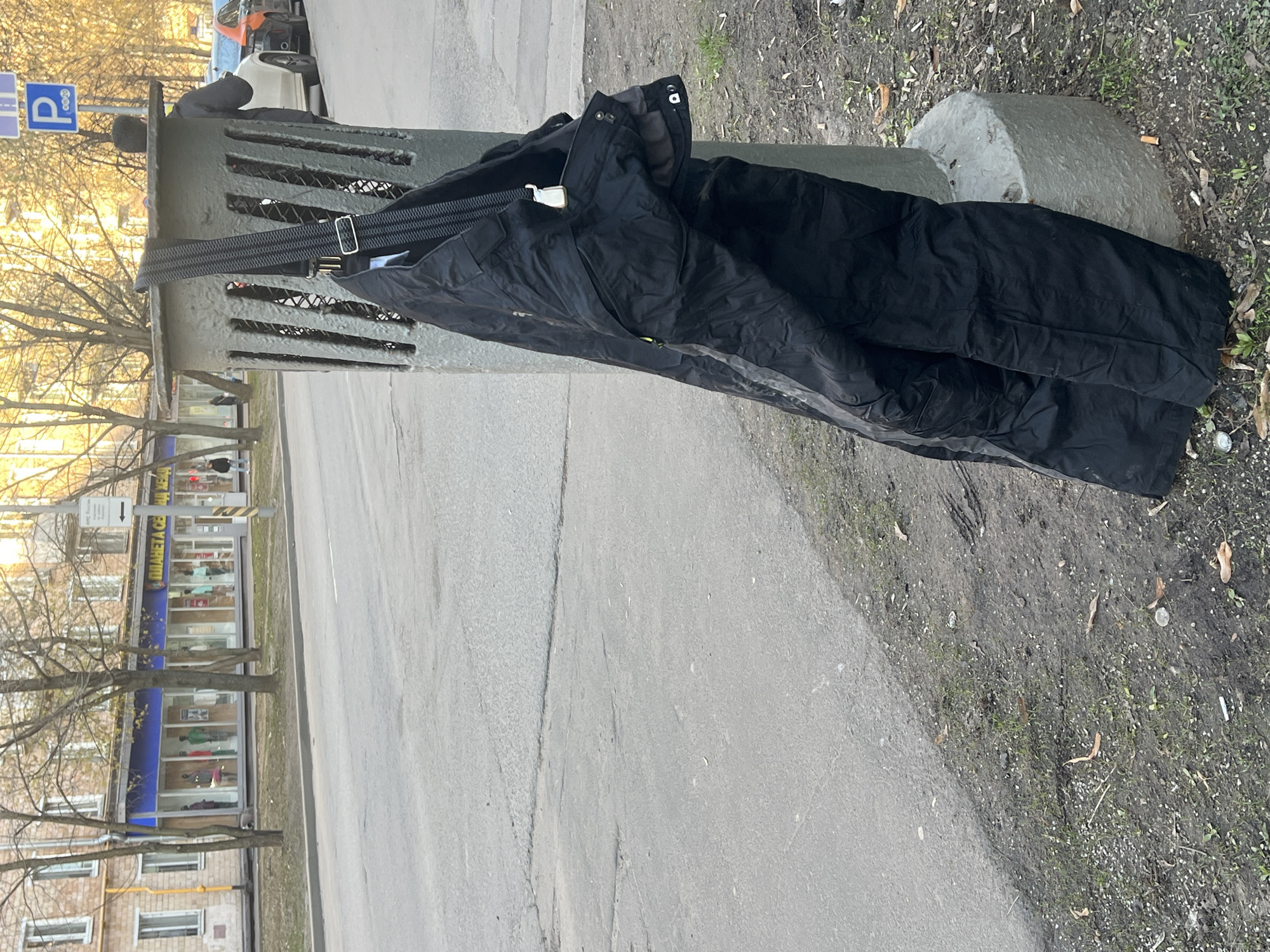 The image displays an outdoor urban street scene, oriented vertically but depicting a horizontal view. The central focus is a pair of black, likely outdoor or ski pants with black suspenders, draped over a grey, slatted concrete or metal barrier. A silver rectangular buckle is visible on the suspenders. To the right of the barrier, a grey, irregularly shaped concrete block rests on the dirt ground.
The setting includes a paved sidewalk and an asphalt road, with a narrow strip of dirt and sparse grass separating them, scattered with fallen leaves and what appears to be a discarded cigarette butt. Bare deciduous trees line the street, suggesting late autumn, winter, or early spring. In the background, a multi-story building with multiple storefronts on its ground level is visible across the street. One shop sign is blue with white Cyrillic text, partially legible as "ДЛЯ ВСЕЙ СЕМЬИ" (FOR THE WHOLE FAMILY).
On the left side of the frame, a blue parking sign with a white "P" and a right-pointing arrow is visible. A person in dark clothing is partially obscured behind the barrier, and another individual can be seen further down the street near the shops.
The lighting indicates either late afternoon or early morning, with warm, soft sunlight illuminating the scene from a low angle. There is no discernible specific activity taking place, other than the presence of the clothing on the barrier, possibly left there temporarily. The location, with its Cyrillic text and urban environment, aligns with the context of Khimki, Russia.
