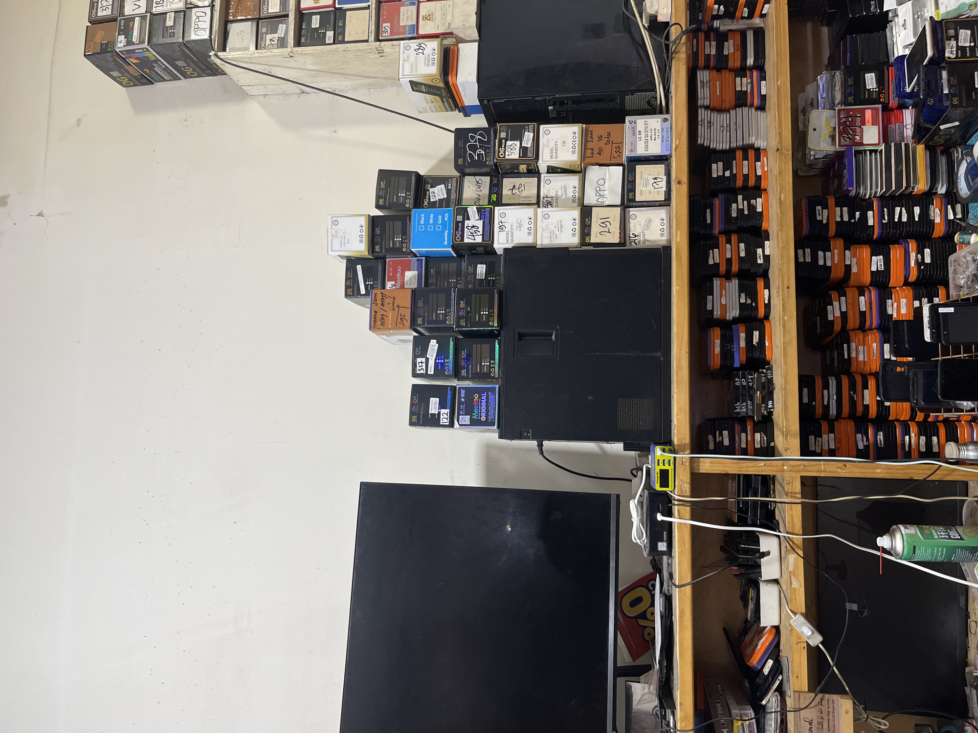The image depicts a cluttered storage or workshop area, likely in a commercial establishment in Central Jakarta, Indonesia. The room is filled with shelves overflowing with various electronic components and accessories, primarily packaged in small boxes and containers. Stacks of boxes, many labeled with brand names like "OG PLUS" and "OPPO," as well as model numbers and quantities, are piled high on top of each other. Some boxes are open, revealing their contents.
In the foreground, a large, dark computer monitor is placed on a desk or table. Cables, chargers, and a can of compressed air are also visible on the desk. A wall-mounted shelf unit holds more stacks of small electronic parts, sorted by color and type. The walls are plain and appear to be made of concrete or plaster, with some signs of wear and tear. The overall impression is one of a busy, functional space dedicated to the repair or sale of electronic devices and their parts. There are no people visible in the image.