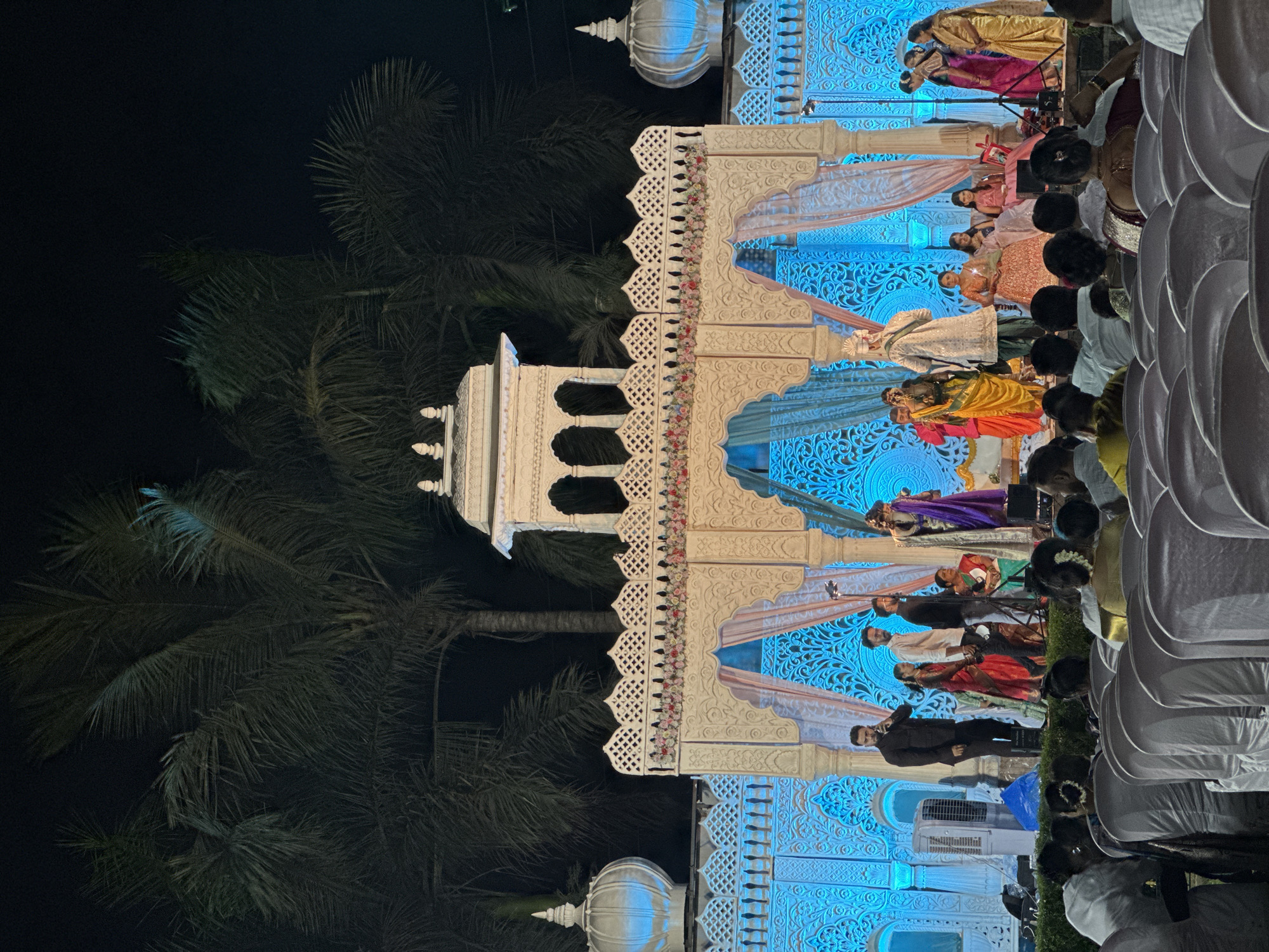 A scene from what appears to be an outdoor wedding or ceremony is captured at night in Shahapur, India. Palm trees sway in the background against the dark sky, beside white architectural structures, including a tower and an ornately decorated platform. The platform is lit up with blue lighting and adorned with flowers, and various individuals are positioned within its sections, indicating different roles or functions in the ceremony. A gathering of people is visible to the side, watching the events unfold, and rows of white chairs are set up for the audience. The setting evokes a sense of traditional festivity.