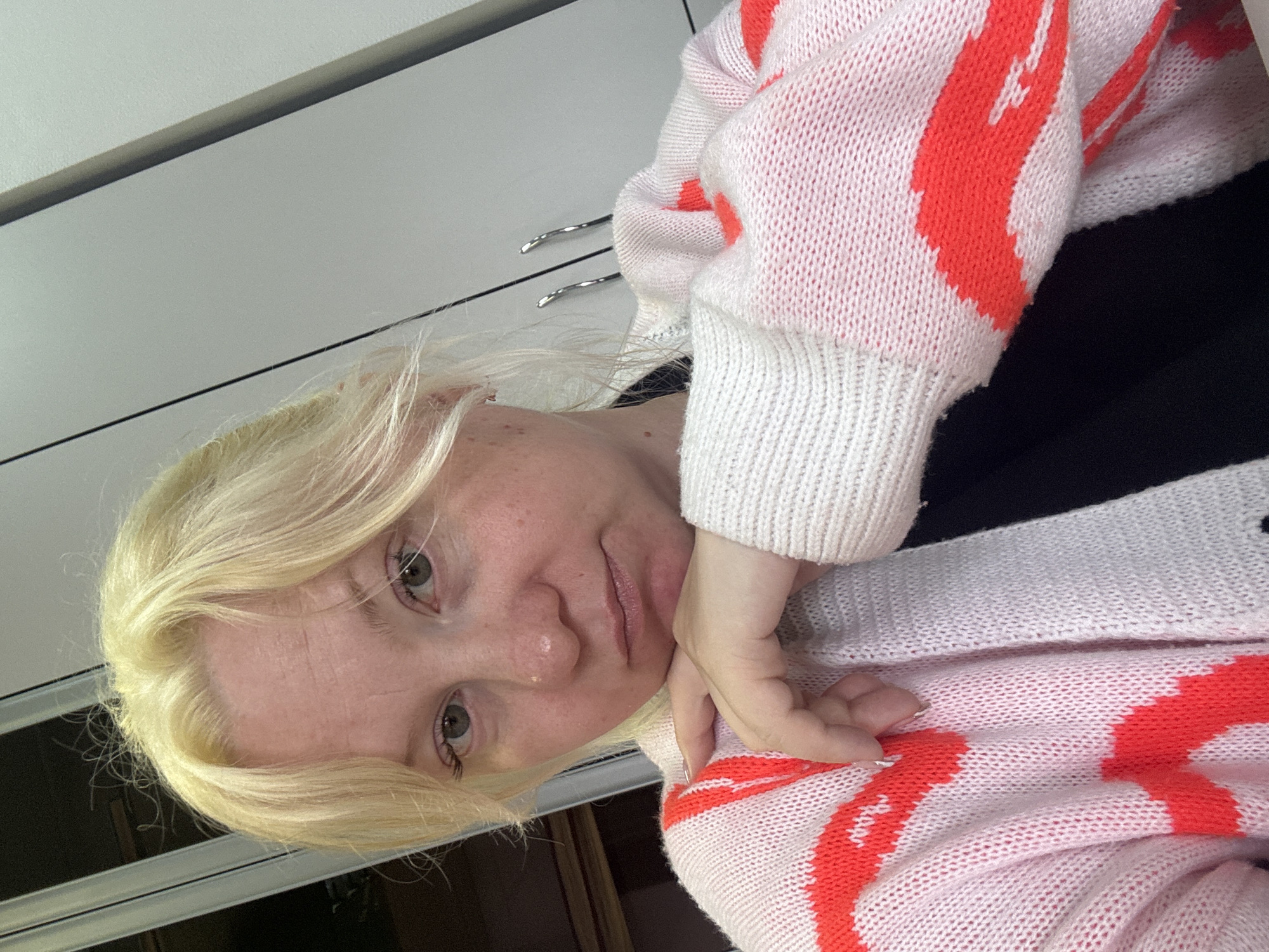 A blonde-haired woman is indoors, possibly in an apartment or house in Chaikovsky, Russia, given the request to specify the location. She is wearing a white and pink knit cardigan with orange abstract patterns, and a black top underneath. Her face is turned to the side, her right hand propped up against her cheek. Part of a white cabinet with silver handles is visible in the background. The image is slightly tilted.