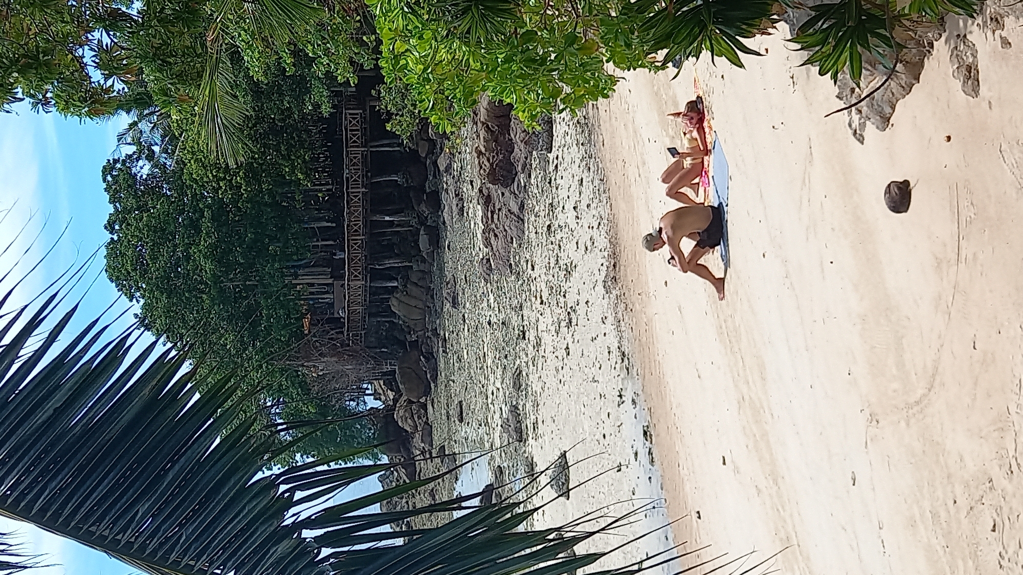 The image captures a beach scene in the subdistrict of Koh Phangan, Thailand. Two individuals are relaxing on the sand. A man is seated on a blue towel, facing away from the camera, and appears to be looking at his phone. A woman is lying on a pink towel next to him, also facing away from the camera, and holding a phone. In the background, a wooden deck structure is visible amongst dense green foliage, with large rocks and the sea beyond. Palm fronds frame the left side of the image. The tide appears to be low, revealing a sandy seabed with scattered rocks and debris.