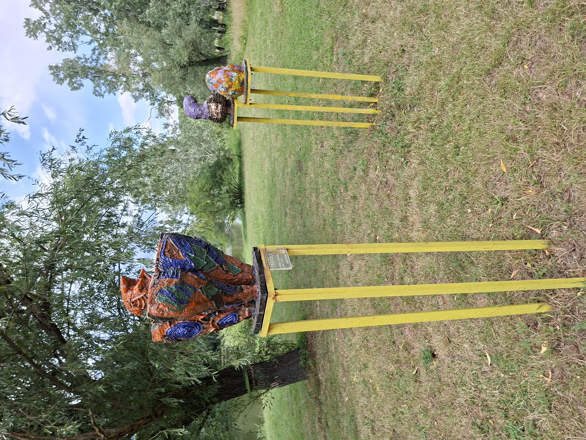 In the city of Chișinău, Moldova, a scene unfolds in an outdoor setting, likely a park or garden. The foreground is dominated by dry, patchy grass, with fallen leaves scattered about. Two yellow, metal stands are prominent. The stand on the left supports a large, intricately designed ceramic sculpture. This sculpture, in shades of orange, blue, and green, appears to be a vessel with a decorative, almost face-like element at its top. A small, rectangular plaque is affixed to the yellow stand, bearing text that likely identifies the artwork and artist. To the right, a second, taller yellow stand holds two smaller ceramic sculptures. The sculpture on the left of this pair is a dark, textured form resembling a bird. Next to it, the sculpture on the right is an egg-shaped object adorned with colorful patterns of orange, yellow, and green. In the background, lush green trees with dense foliage line a body of water, possibly a lake or river. The sky above is a bright blue, dotted with white clouds. The overall impression is of an art installation in a natural environment.