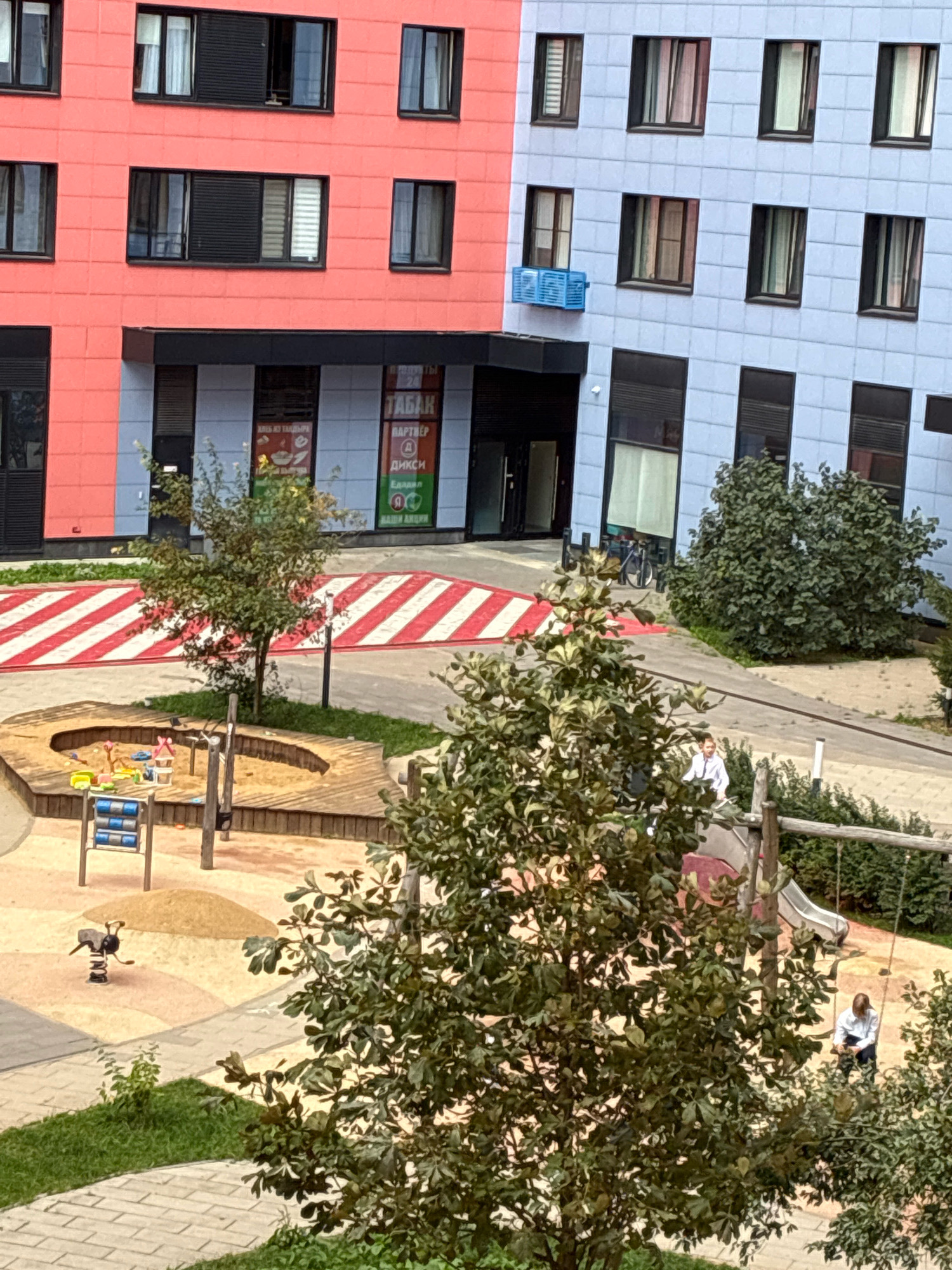The image depicts an exterior view of a modern residential building in the city of Kommunarka, Russia. The building features a striking facade with sections of coral pink and light blue. The lower floors of the building house commercial spaces with glass entrances. In front of the building, there is a children's playground with a sandbox, a spring rider, and a swing set with a slide. A red and white striped crosswalk is visible on the paved ground. Trees and greenery are interspersed throughout the scene. Two individuals are present: one person is seated on a swing set on the playground, and another person is standing near a bicycle in the background. The lighting suggests it is daytime.
