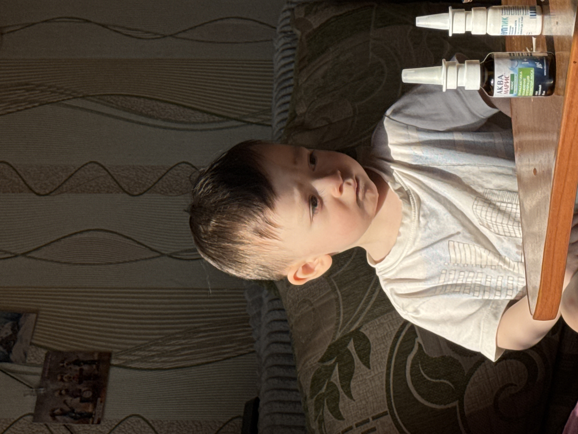 A young child is sitting next to a small wooden table. On the table are two nasal spray bottles: "Аква Марис" and "Ринолайк." The scene is indoors, with a patterned wallpaper and furniture in the background. The location is Izhevsk, Russia. A picture frame is also visible on a table to the bottom-left of the image.