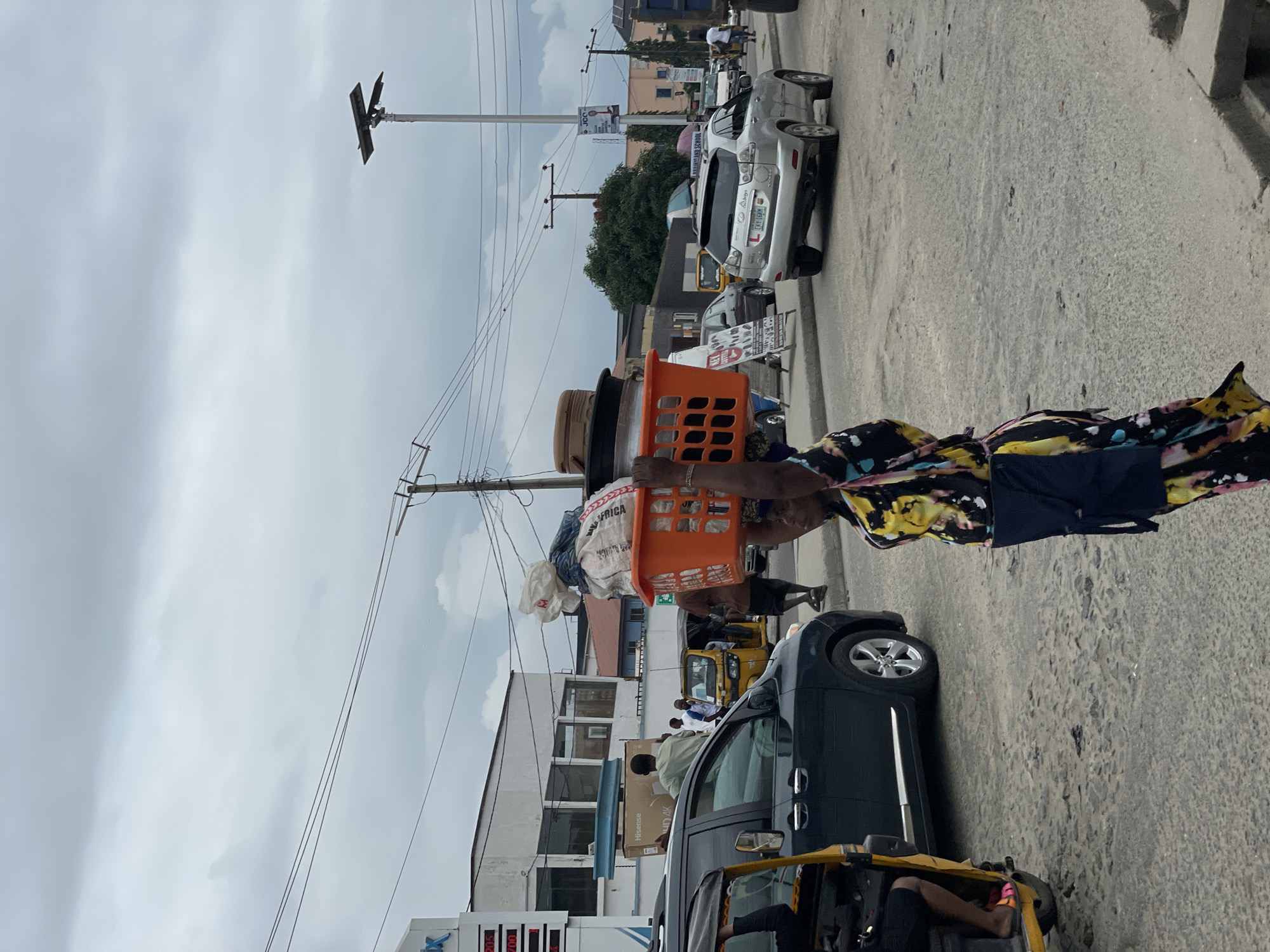 A woman walks with a large orange plastic basket filled with goods balanced on her head in a bustling street scene in Ado, Nigeria. The basket contains folded textiles and what appears to be a large pot lid. She is wearing a colorful patterned dress and a dark blue garment around her waist. In the background, a silver SUV is driving away, its license plate visible. A dark colored minivan is parked on the left, and a yellow tricycle-style vehicle is partially visible in front of it. Other pedestrians, vehicles, and buildings line the street under an overcast sky. The road surface is asphalt with visible patches and cracks. Overhead power lines crisscross the sky, and a street lamp with solar panels is prominent. The overall impression is a snapshot of everyday urban life and commerce.