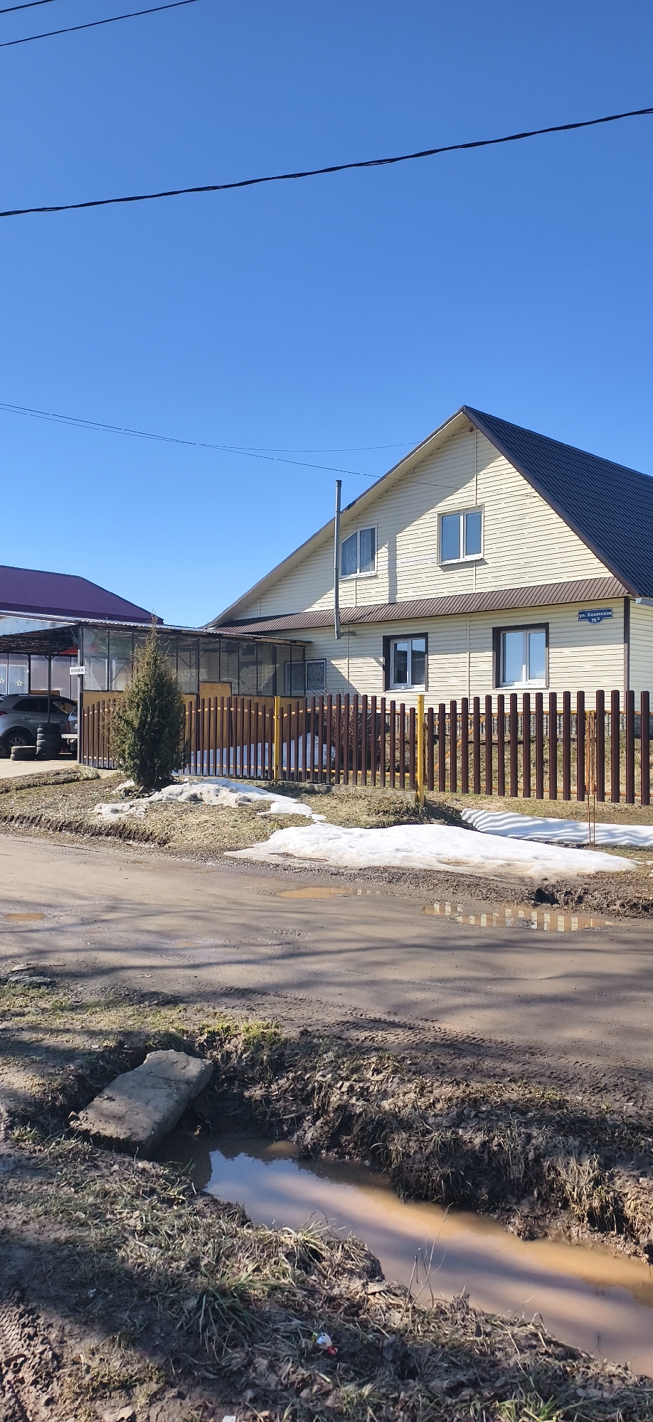 A medium shot depicts a muddy street scene in Фоки, Russia. A two-story house is visible in the background, featuring a cream-colored exterior, brown trim around the windows, and a brown roof. A sign indicating the street name, "ул. Советской 76 а," is attached to the side of the house. A brown wooden fence with yellow posts borders the property, separating it from the street. Patches of melting snow are scattered on the ground near the fence. To the left of the house is a structure with a purple roof, a dark vehicle, and a stack of tires. The foreground is dominated by a dirt and mud road, with a ditch running alongside it. A flat concrete slab serves as a makeshift bridge over the ditch. The sky is a clear, bright blue, with some electrical wires visible.