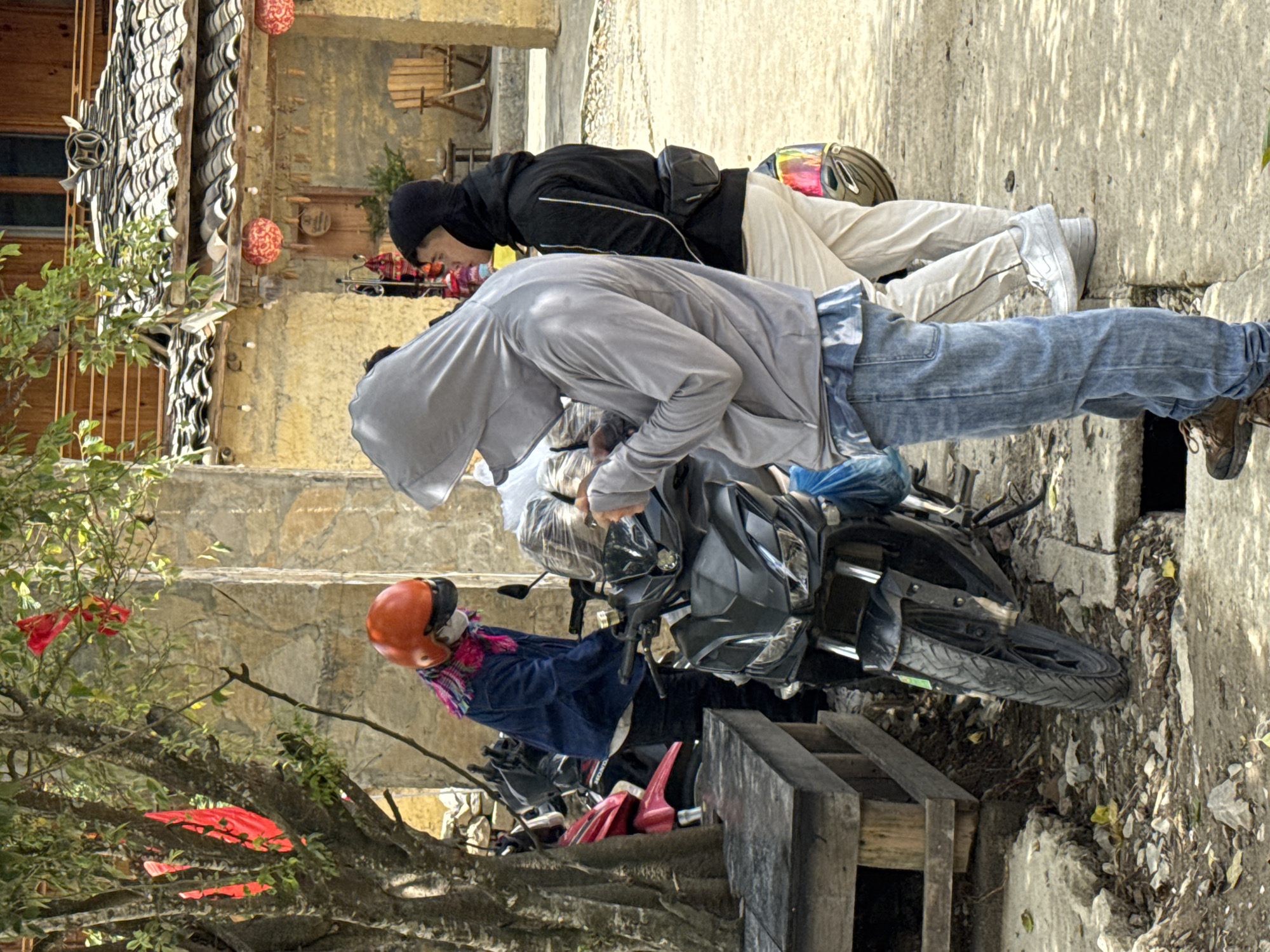 Two people are bent over looking at items for sale. One person is wearing a gray hooded sweatshirt, jeans, and brown shoes. The other person is wearing a black hooded jacket, white pants, and white shoes. Two motorcycles are parked nearby. One motorcycle is black and the other is red and pink. The location appears to be in a street in Lũng Cú Commune, Vietnam. The environment includes buildings with tile roofs and a stone wall. A tree is also present.