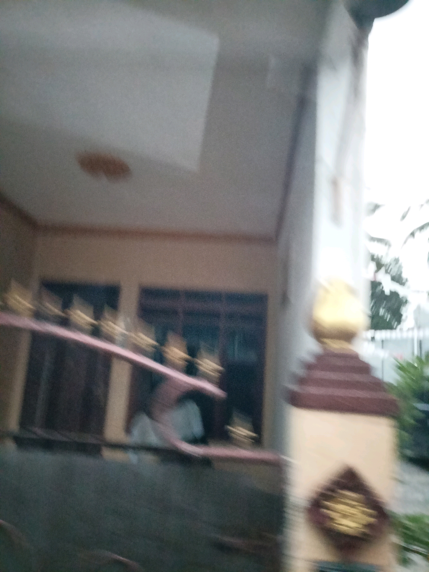 The image, taken in Payaman, Indonesia, is an out-of-focus shot of the exterior of a building. A prominent architectural feature is a red, stepped pillar topped with a golden, bulbous ornament. The building itself appears to have light-colored walls and dark-framed windows. In the foreground, a pink railing is visible, with a hint of dark wood or metal underneath it. The scene suggests a residential or commercial property with some decorative elements. The overall lighting is bright, indicating it was taken during the daytime.
