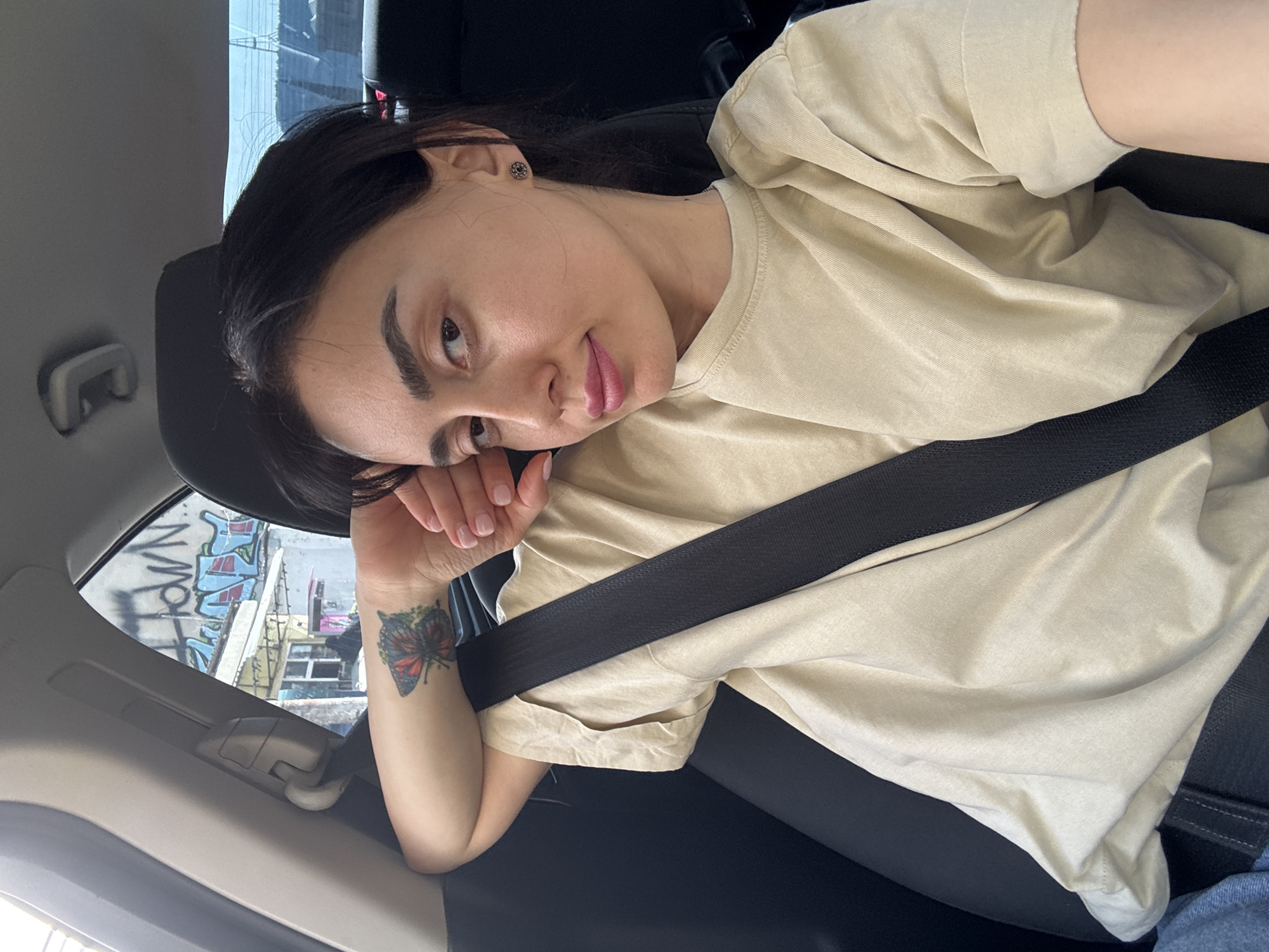 A woman is reclining in the passenger seat of a car. She is wearing a beige t-shirt and a seatbelt. Her left hand is resting on her face and her arm bears a butterfly tattoo. Through the car window, buildings with graffiti can be seen. The graffiti and architecture are consistent with the environment of Tbilisi, Georgia.