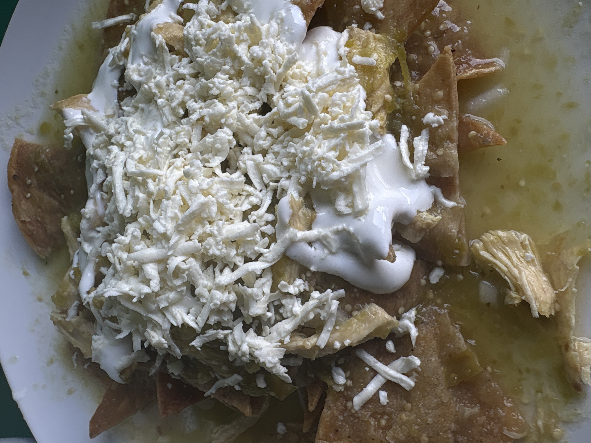 A close-up shot of a plate of chilaquiles, a traditional Mexican dish, likely taken in Mexico City, Mexico. The chilaquiles are bathed in a green salsa, topped with crumbled cheese and a dollop of cream. Tortilla chips are visible beneath the toppings and sauce. The dish is served on a white plate. The scene appears to be indoors, potentially in a home or restaurant setting.