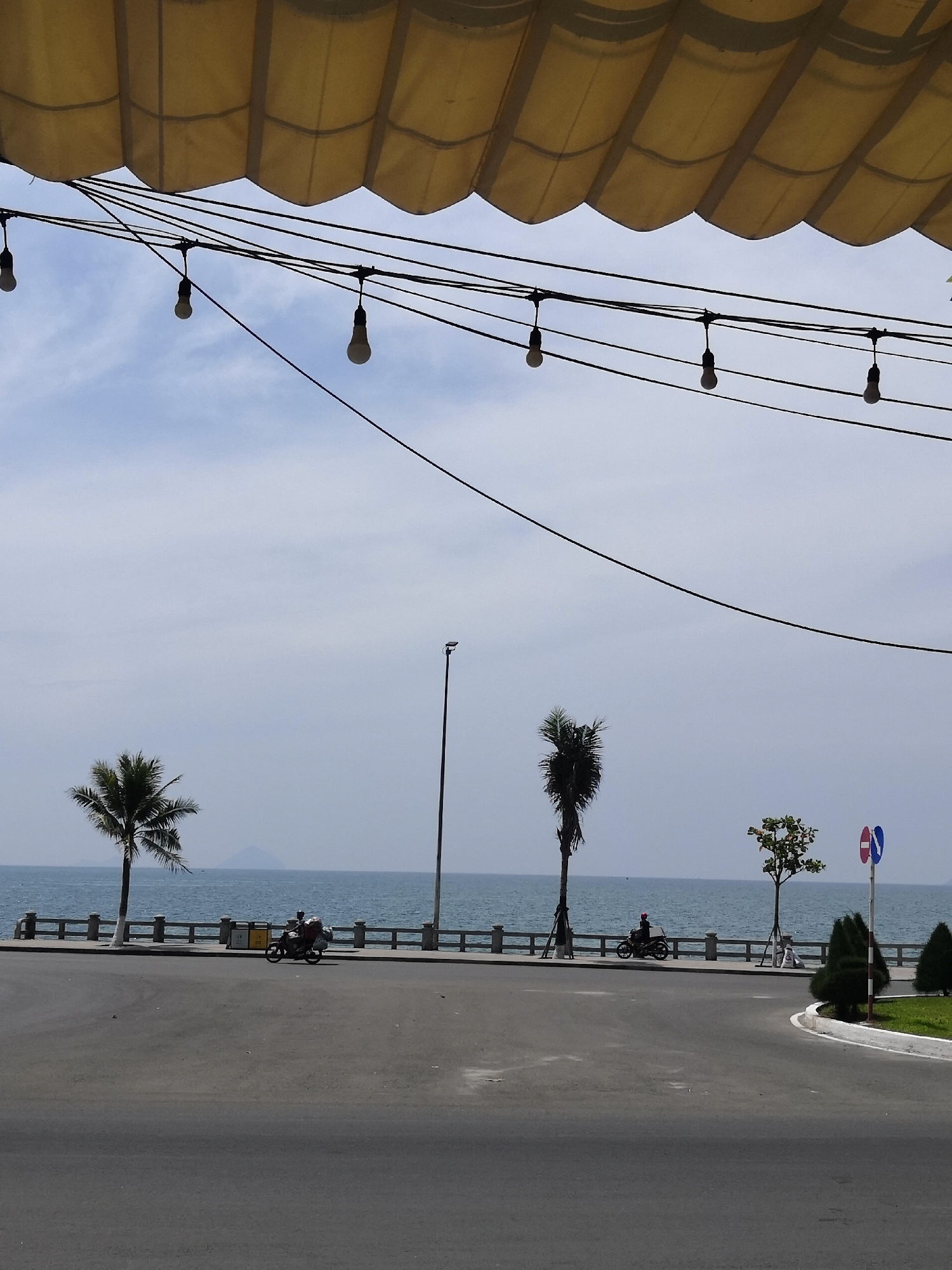 A wide shot captures a street scene facing the ocean, with a prominent yellow awning overhead and strings of bare lightbulbs hanging across the frame. The foreground is dominated by an asphalt road. Behind it, a paved walkway runs parallel to a low concrete barrier. Beyond the barrier, the blue ocean stretches to the horizon, with a distant, hazy mountain visible on the left. Two palm trees stand on the left side of the image, with a single motorcycle carrying a rider and cargo seen on the walkway. Further to the right, another, thinner palm tree and a small leafy tree are present. A lamppost stands near the center, and a traffic sign with blue and red circles is visible on the right. The sky is a pale, hazy blue with light clouds. The scene is likely in Bac Nha Trang Ward, Vietnam, given the tropical vegetation and coastal setting.