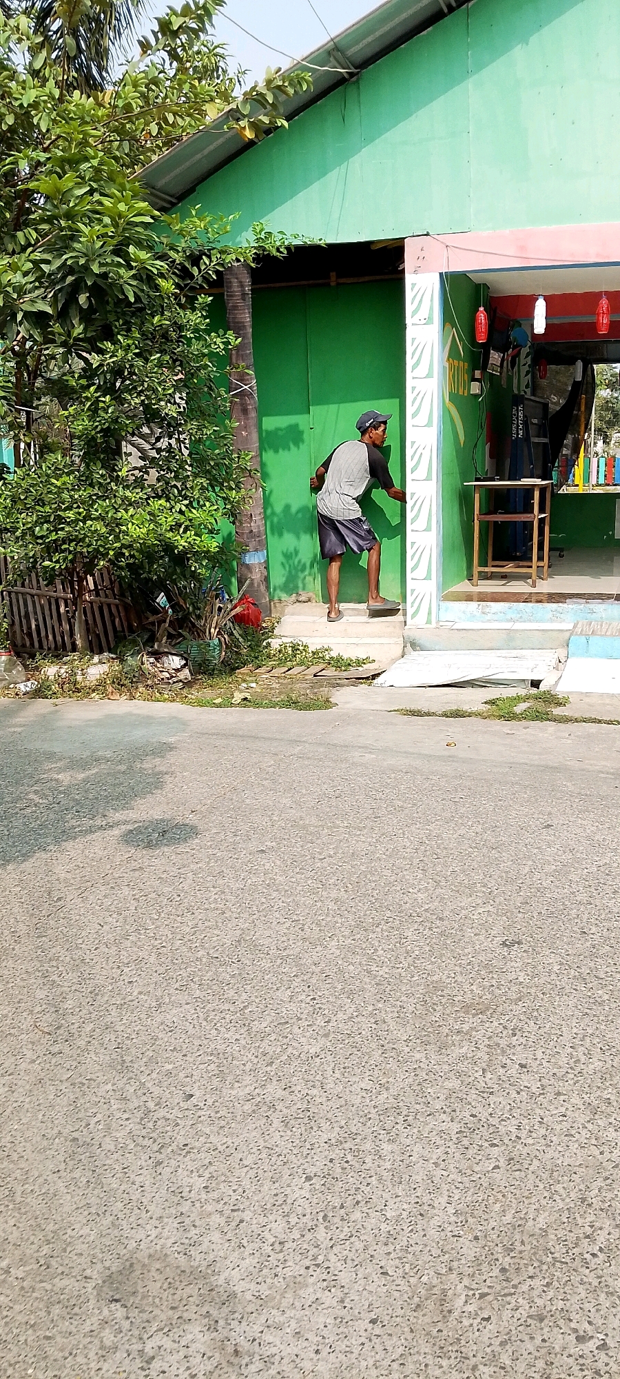 A man wearing a dark baseball cap, a grey t-shirt with black sleeves, and dark shorts is standing on a white staircase outside a green building. He is facing away from the camera, with his right hand on the green wall. The building appears to be a shop, with a red and white awning above the entrance, and shelves stocked with goods visible inside. The man is on the second step from the bottom of a short set of stairs. In front of the building is a paved area, and beyond that is a road. To the left of the building, there are trees and vegetation, and a wooden fence. The overall scene is outdoors, under a bright sky, suggesting a daytime setting. The location is identified as Satriamekar, Indonesia.