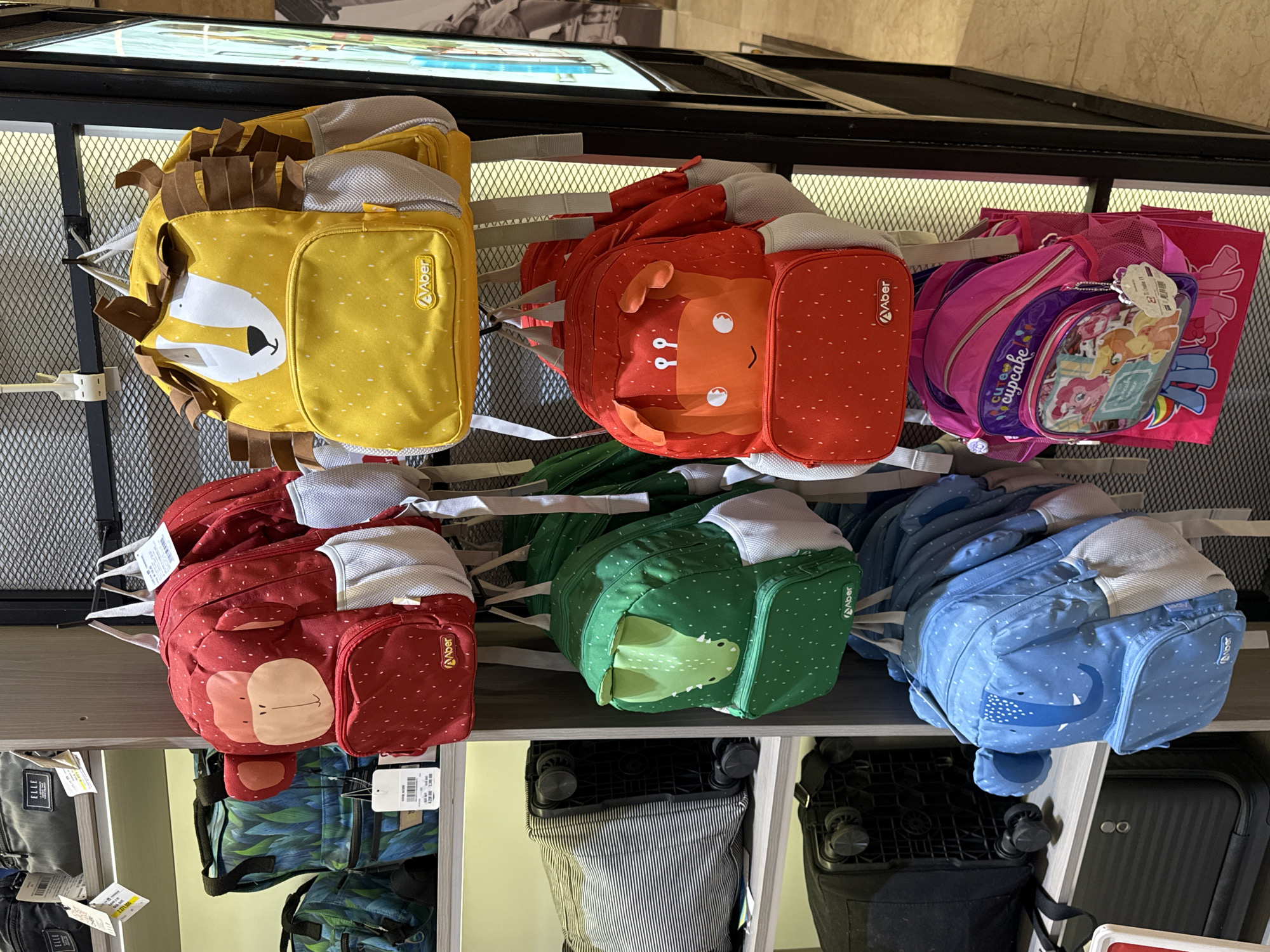 The image depicts a retail display of children's backpacks in a store. The backpacks are arranged on shelves and hung from hooks. Several backpacks are designed with animal themes, including a red monkey, a yellow lion, a green alligator, and a blue elephant. There is also a pink backpack with a "cupcake" design and characters from My Little Pony. In the background, there are other bags and luggage items on shelves. The store appears to be well-lit and organized. The location is identified as a store in Nha Trang Ward, Vietnam, suggesting it is a commercial establishment within that city. There are no people visible in the image.