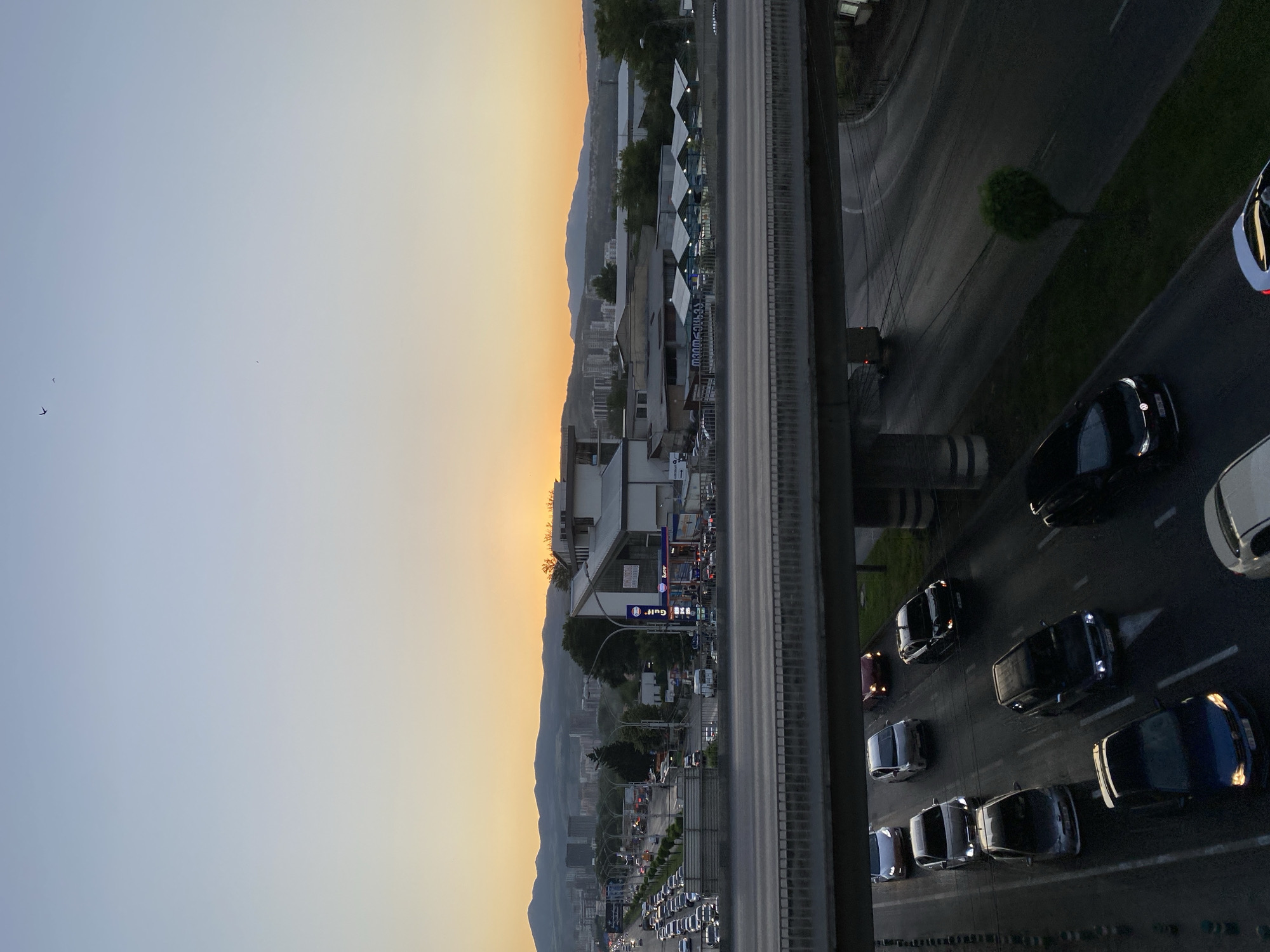 This is a photograph taken from an elevated perspective, likely a building or bridge, overlooking a multi-lane highway in Tbilisi, Georgia. The scene captures a sunset or sunrise, with a gradient of soft orange and blue hues dominating the sky on the left side of the frame. Silhouetted against this colorful sky are distant mountains and the urban landscape of Tbilisi, characterized by numerous buildings and a highway lined with trees.
The highway is filled with cars, indicating moderate to heavy traffic. The vehicles appear to be a mix of sedans and SUVs, traveling in both directions. Some vehicles have their headlights on, suggesting it's either early morning or late evening. The road surface is asphalt, and traffic lanes are clearly marked. On the right side of the highway, there is a grassy verge and a dense, rounded tree.
Notable details include several birds flying in the sky, adding a sense of life to the tranquil sky. There is a building with a distinctive geometric roofline situated along the highway, which appears to be a commercial establishment. A sign on this building displays the "Gulf" logo, indicating a gas station. Other signs, though partially obscured or illegible from this distance, suggest other businesses. The overall mood of the image is peaceful, with the warm glow of the sky contrasting with the busy urban environment. The lighting suggests the golden hour, shortly after sunrise or before sunset.