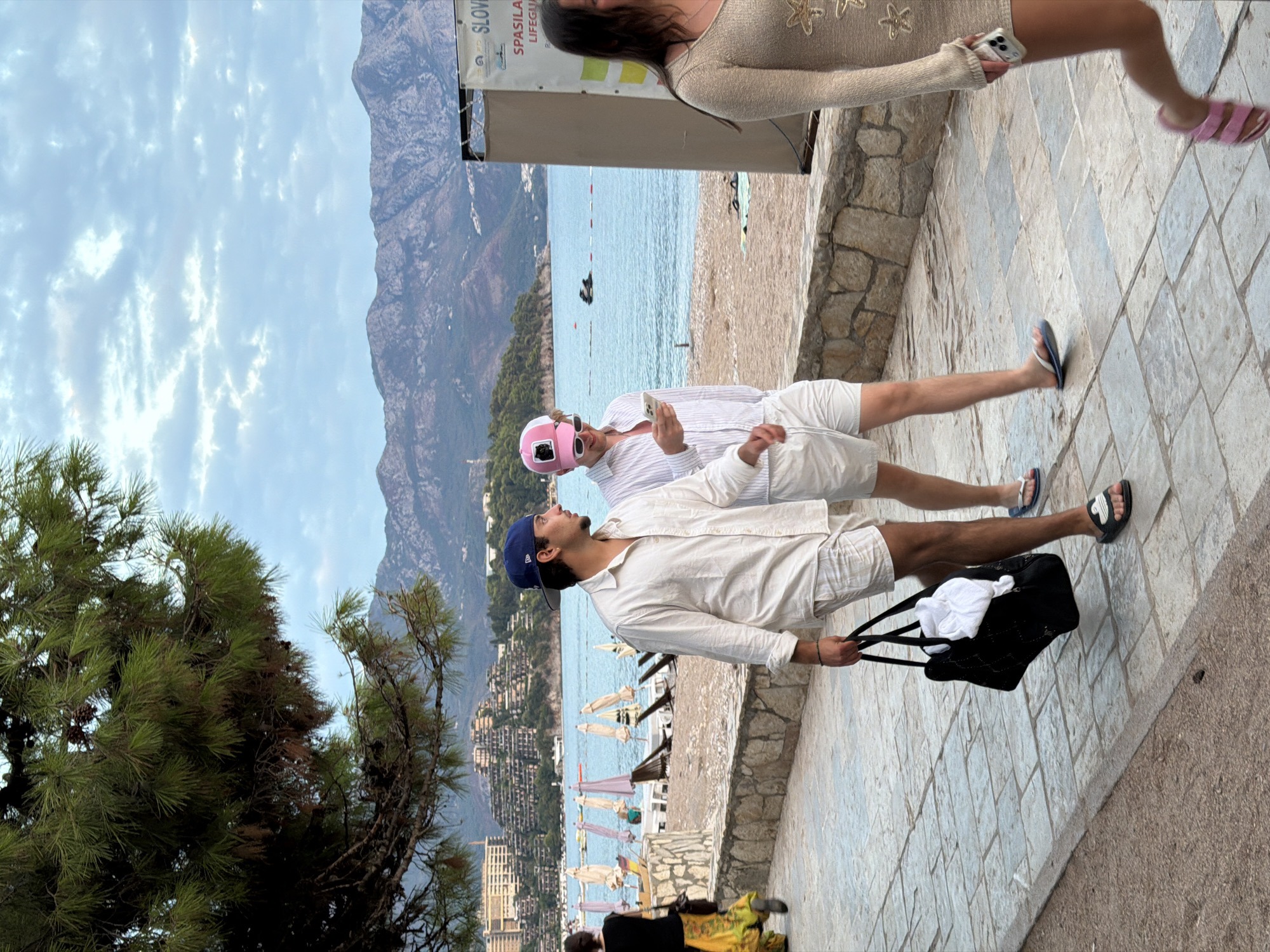 The image depicts a scene on a paved walkway by a beach, with the Adriatic Sea and mountains in the background, likely in Budva, Montenegro. Two young men are walking towards the right side of the frame. The man on the left wears a white linen shirt, white shorts, a blue baseball cap, and black slides. He is carrying a black duffel bag with a white towel protruding from it. The man on the right wears a striped button-down shirt, white shorts, white-rimmed sunglasses, and pink flip-flops. He is looking down at his phone. Behind them, a woman in a long, flowing yellow skirt and black top is walking away from the camera. Further in the background, beach umbrellas and lounge chairs are visible on a pebble beach, and buildings are nestled into the hillside. A sign with "SLOVENIA" and "SPASILA LIFEGU" is partially visible on a booth to the right. A woman in a light-colored, long-sleeved dress with star embellishments is walking towards the left on the far right side of the frame, holding a smartphone. A jet ski is visible in the water. The sky is overcast with scattered clouds.